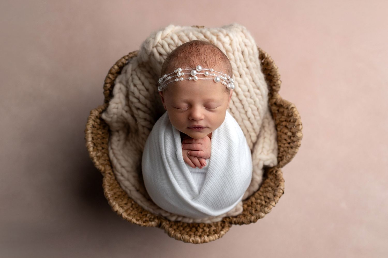 Adorable newborn baby wrapped in white swaddle blanket sleeping in brown knitted basket.