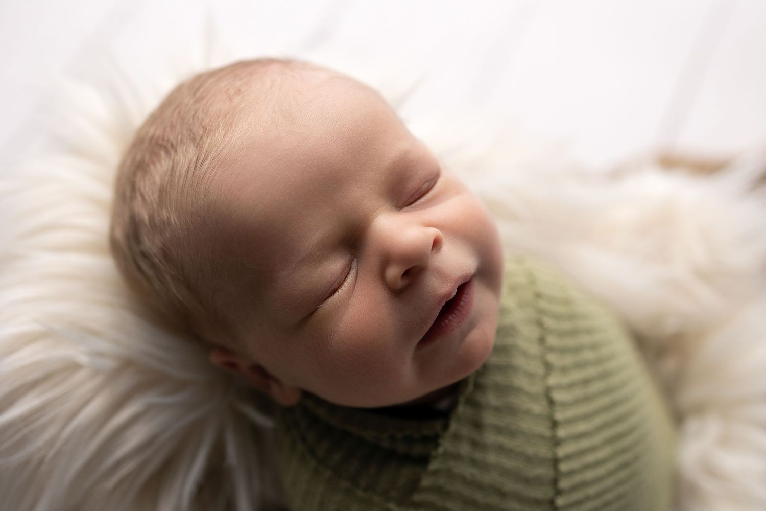 A sleeping baby wearing a green sweater lies peacefully on white bedding.