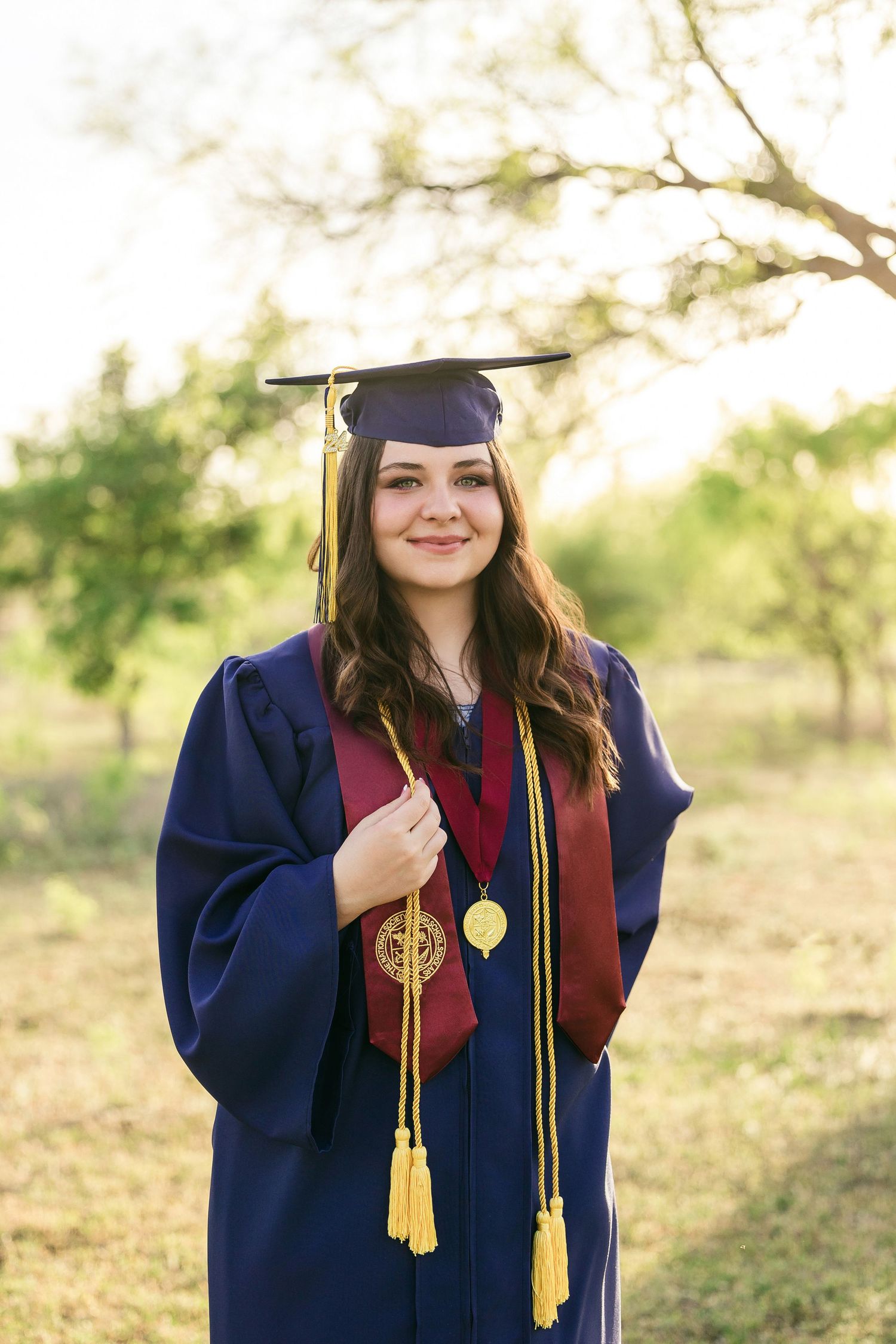 Graduate in navy cap and gown with burgundy stole and gold tassel poses outdoors during sunset photo session.