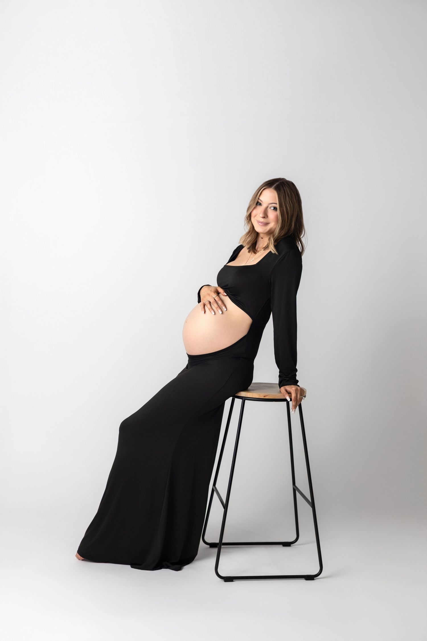 A series of maternity photos showing a pregnant woman in a black dress posing elegantly with a modern stool against white backdrop.