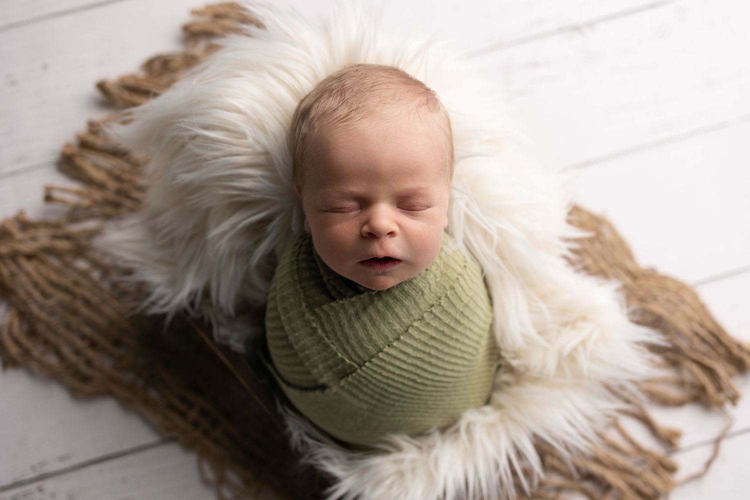A sleeping newborn swaddled in green fabric lies on a soft white fur blanket against a light wooden backdrop.