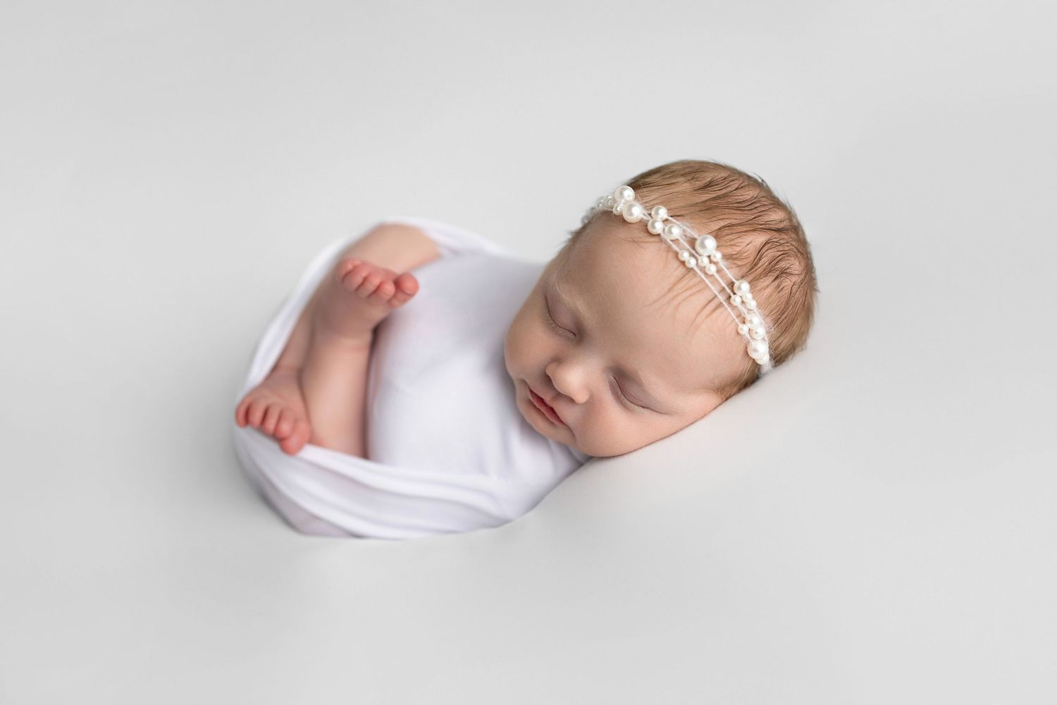 A sleeping newborn baby wearing a white headband lies peacefully on soft white fabric in a minimalist photography setting.