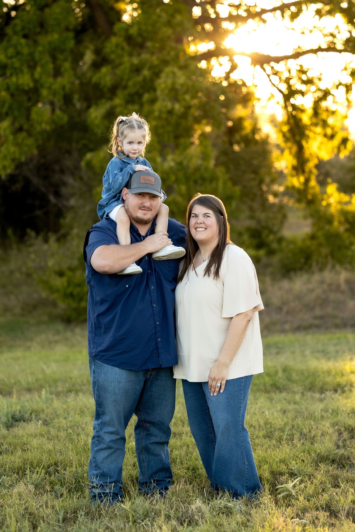 A family stands together in a grassy field at sunset with golden light filtering through trees in the background.