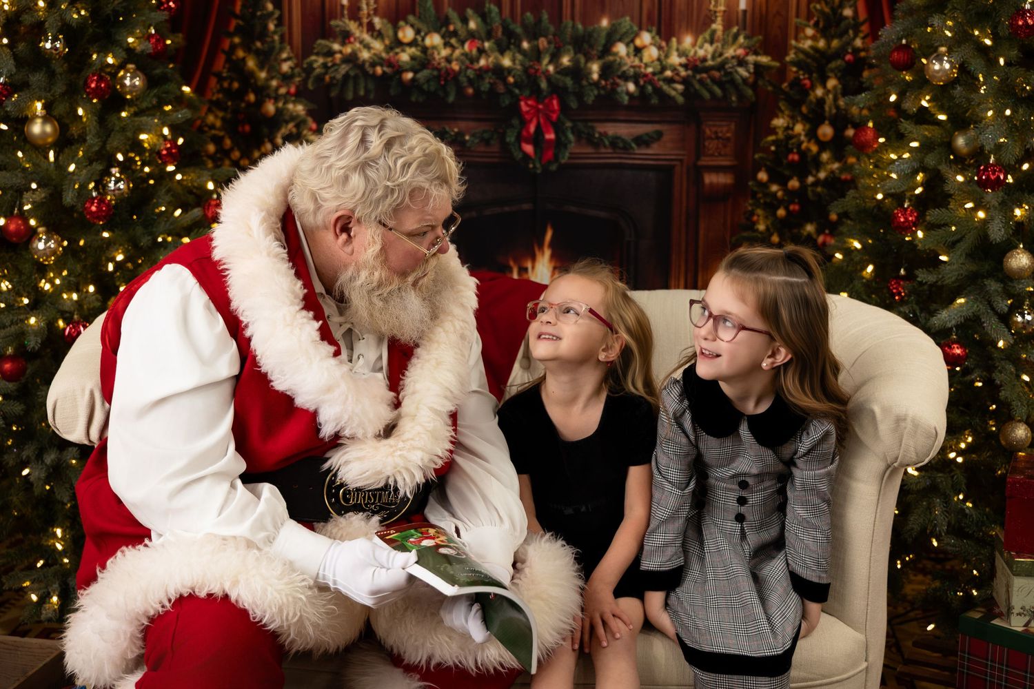 Santa Claus reading his list to two young girls beside decorated Christmas trees and a glowing fireplace.