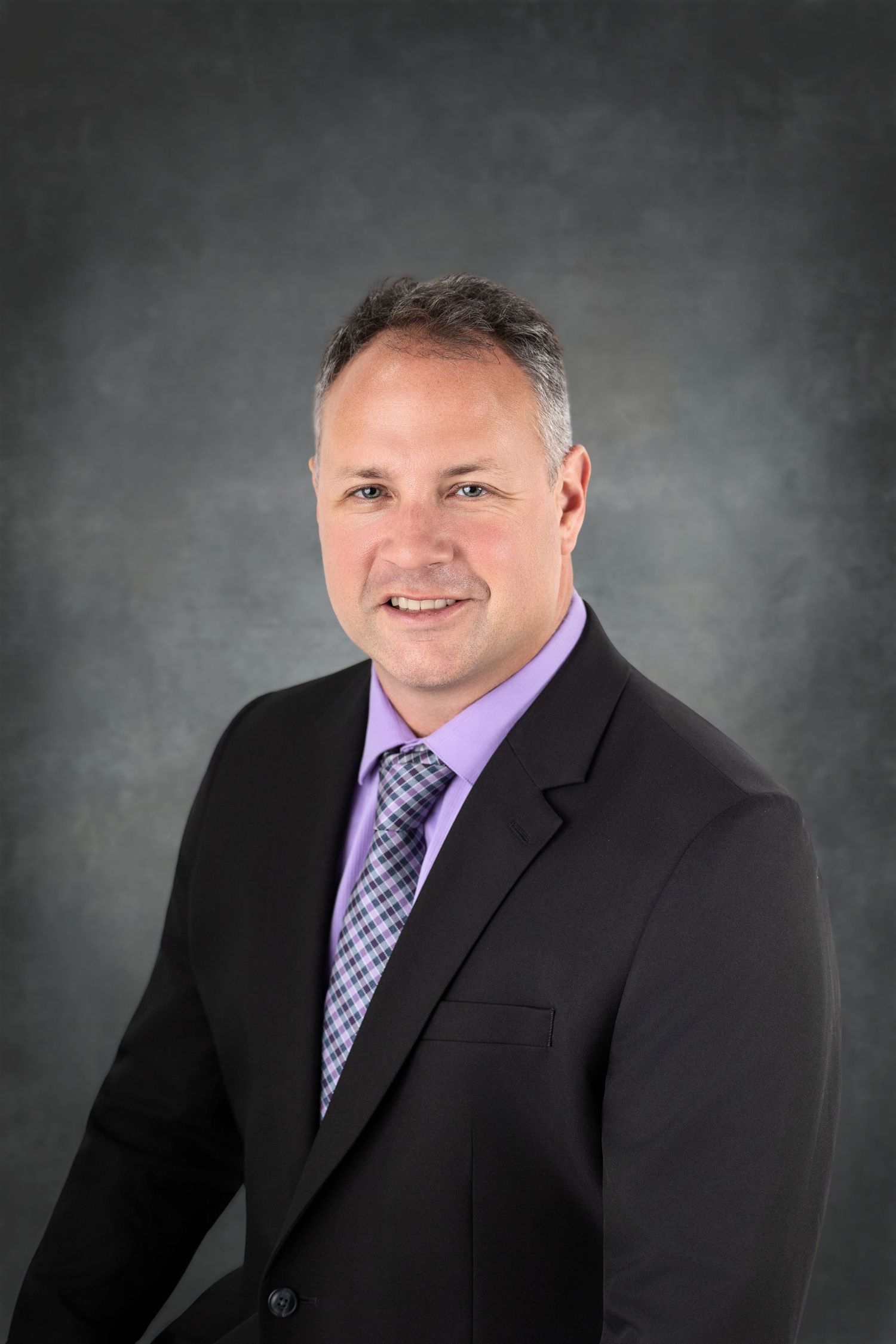 Professional headshot of someone in a black suit with purple striped tie against a dark gray backdrop.