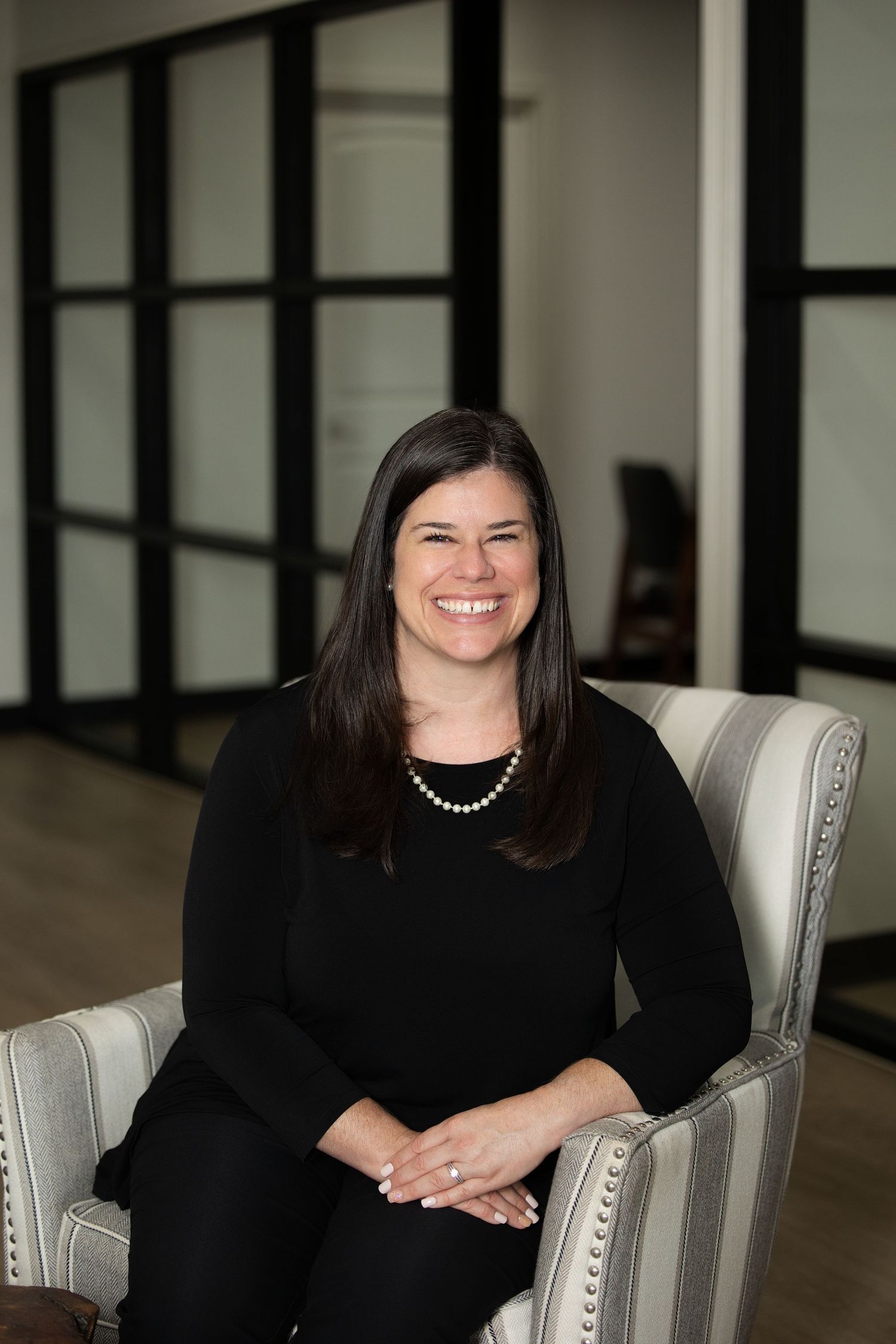 Professional portrait of someone in black attire seated in an elegant gray chair with nailhead trim.