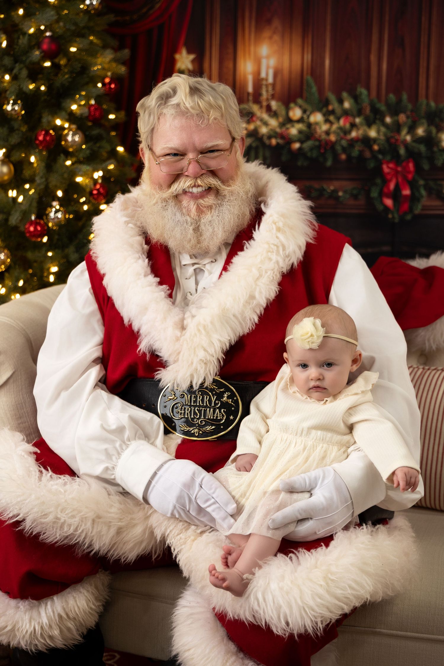 Baby in cream dress sits on Santa Claus lap beside decorated Christmas tree holding Merry Christmas sign.