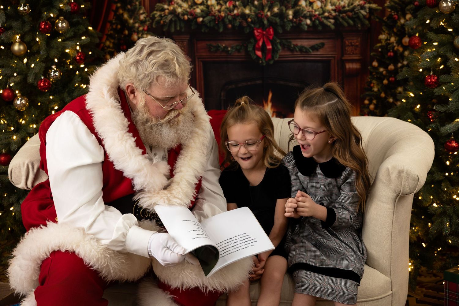 Santa Claus reads a book with two smiling girls on a festive chair surrounded by decorated Christmas trees.