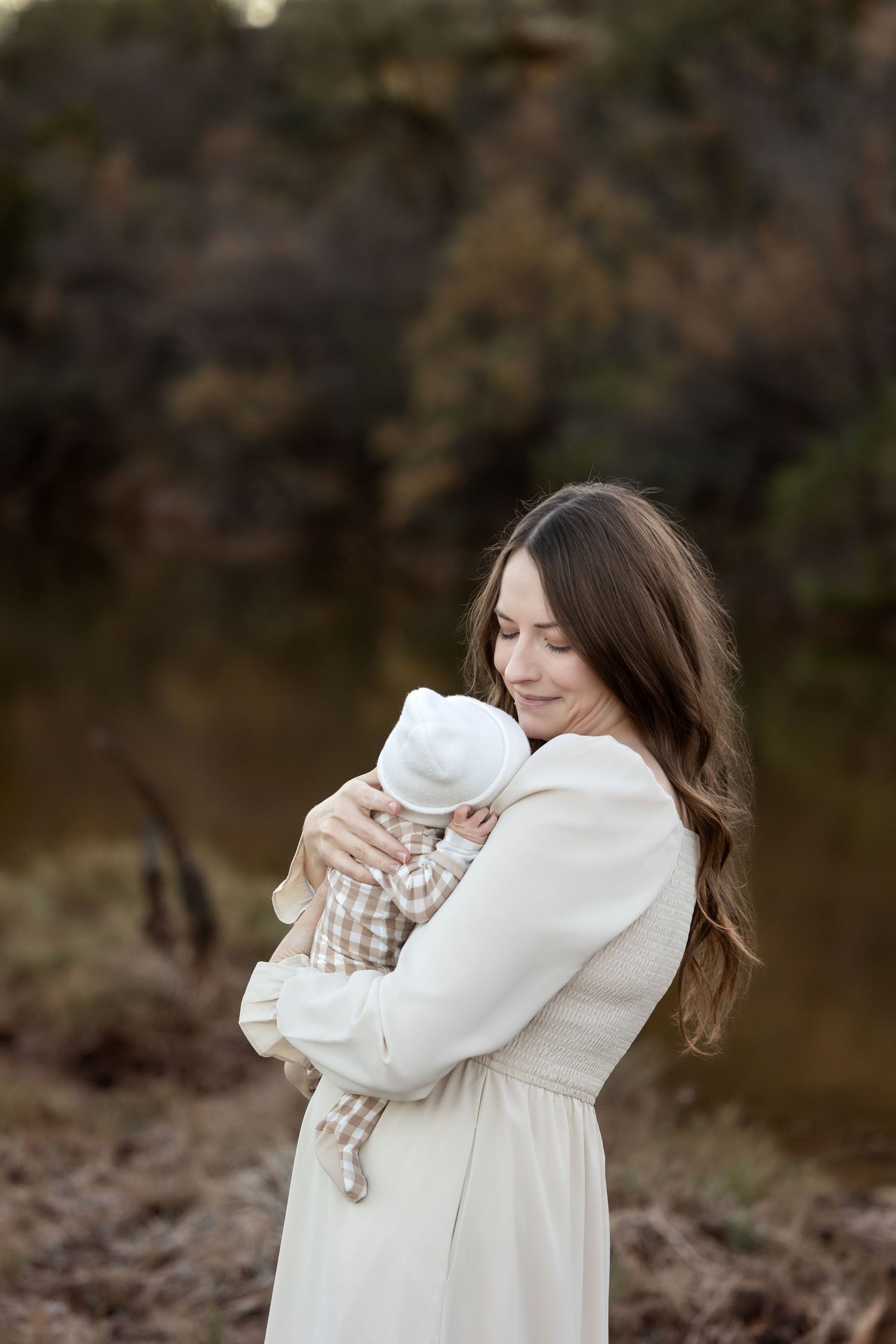 A person in a white dress holds and embraces a small baby wrapped in white blankets outdoors.