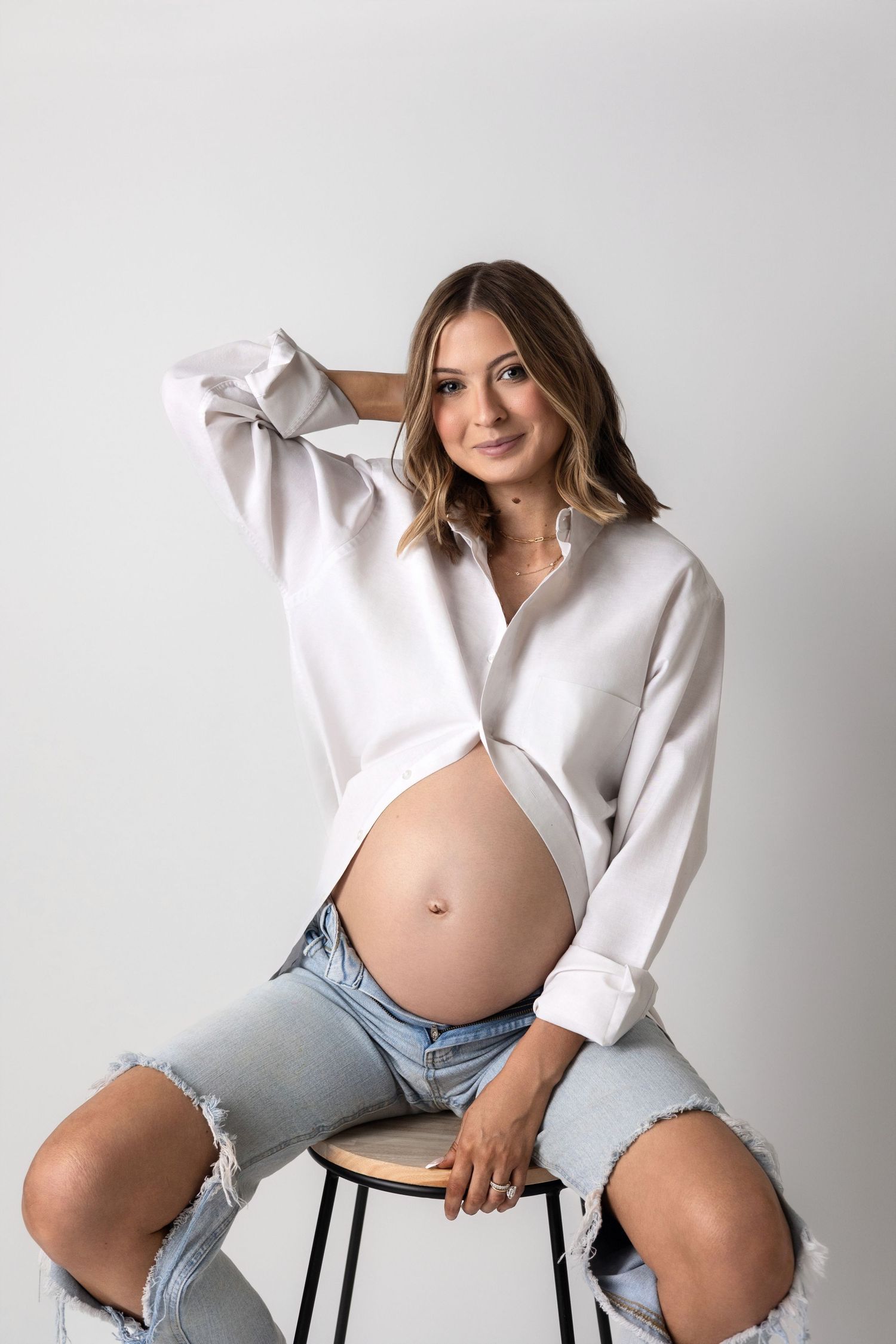 Maternity photoshoot featuring a white button-down shirt and denim shorts against a light gray backdrop.
