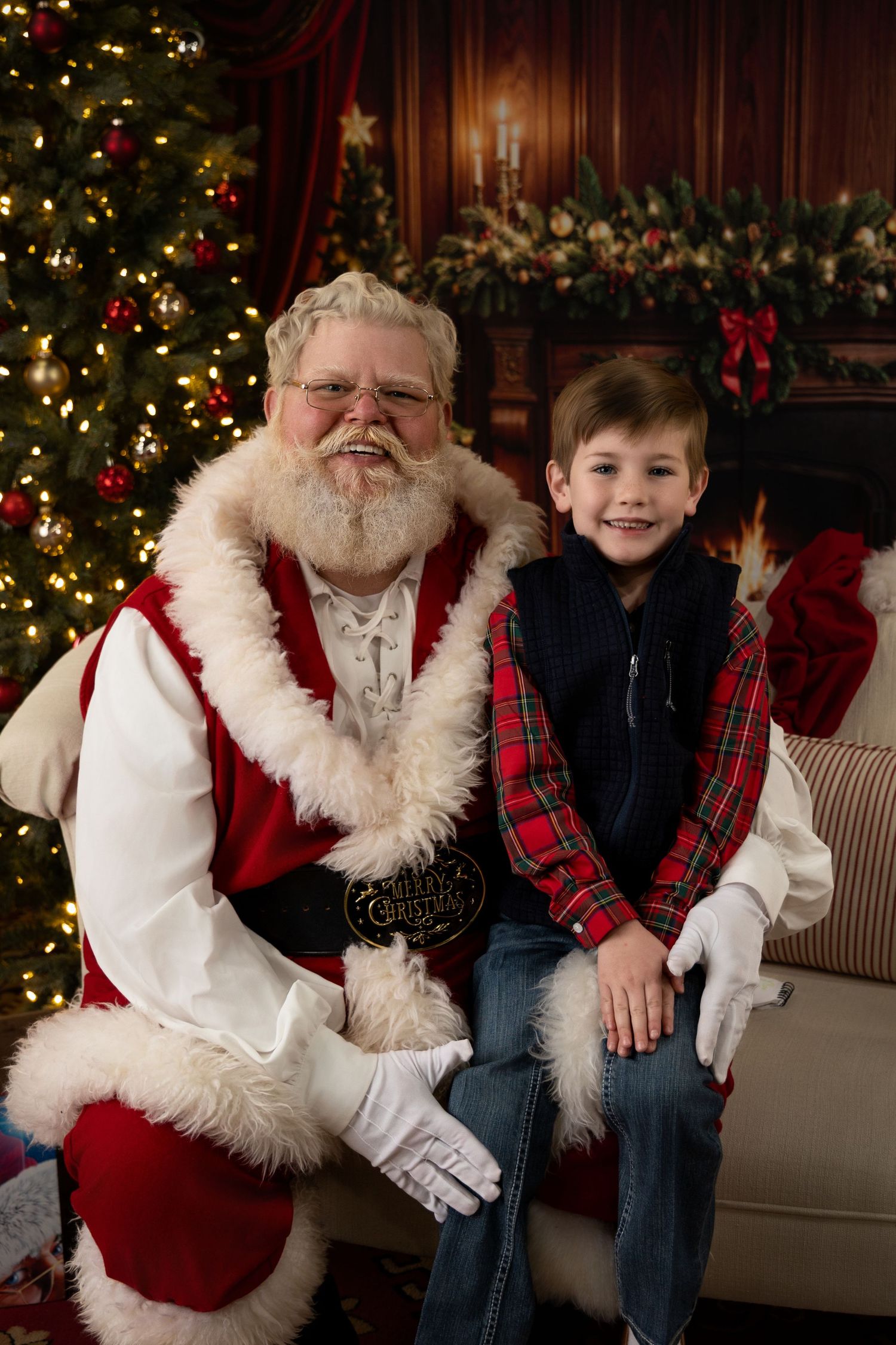 A smiling boy in plaid shirt sits with Santa Claus beside a decorated Christmas tree in a cozy holiday setting.