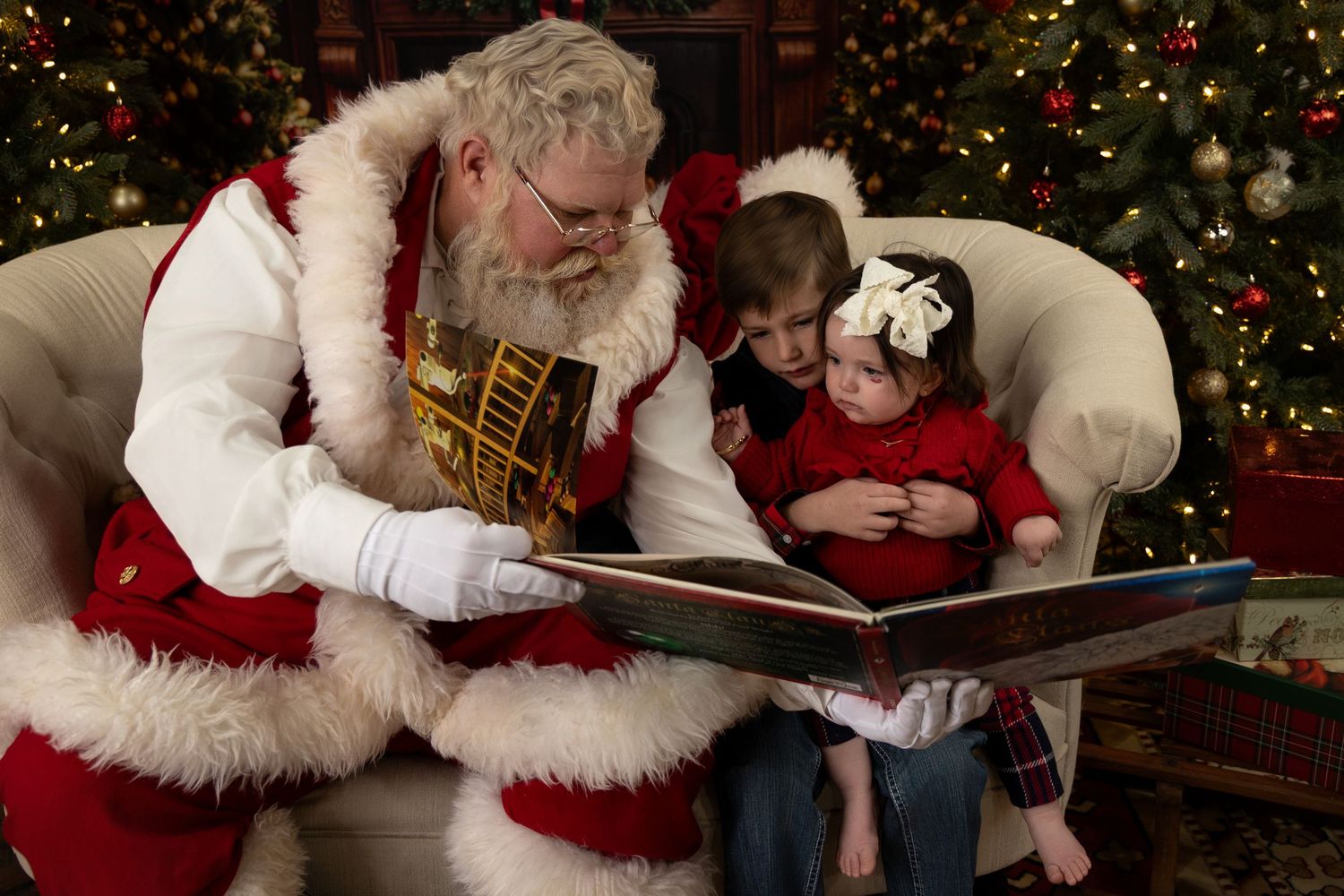 Santa Claus reads a Christmas book to two young children while sitting in a white armchair near a decorated Christmas tree.