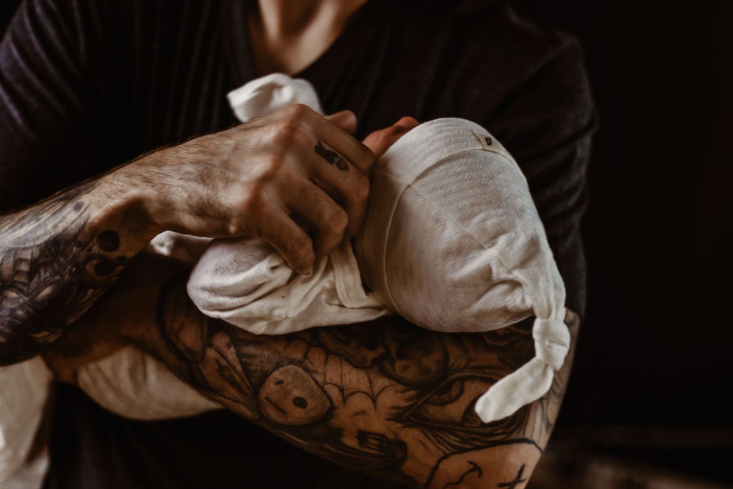A person holding a sleeping newborn baby, with tattoos visible on the forearms.