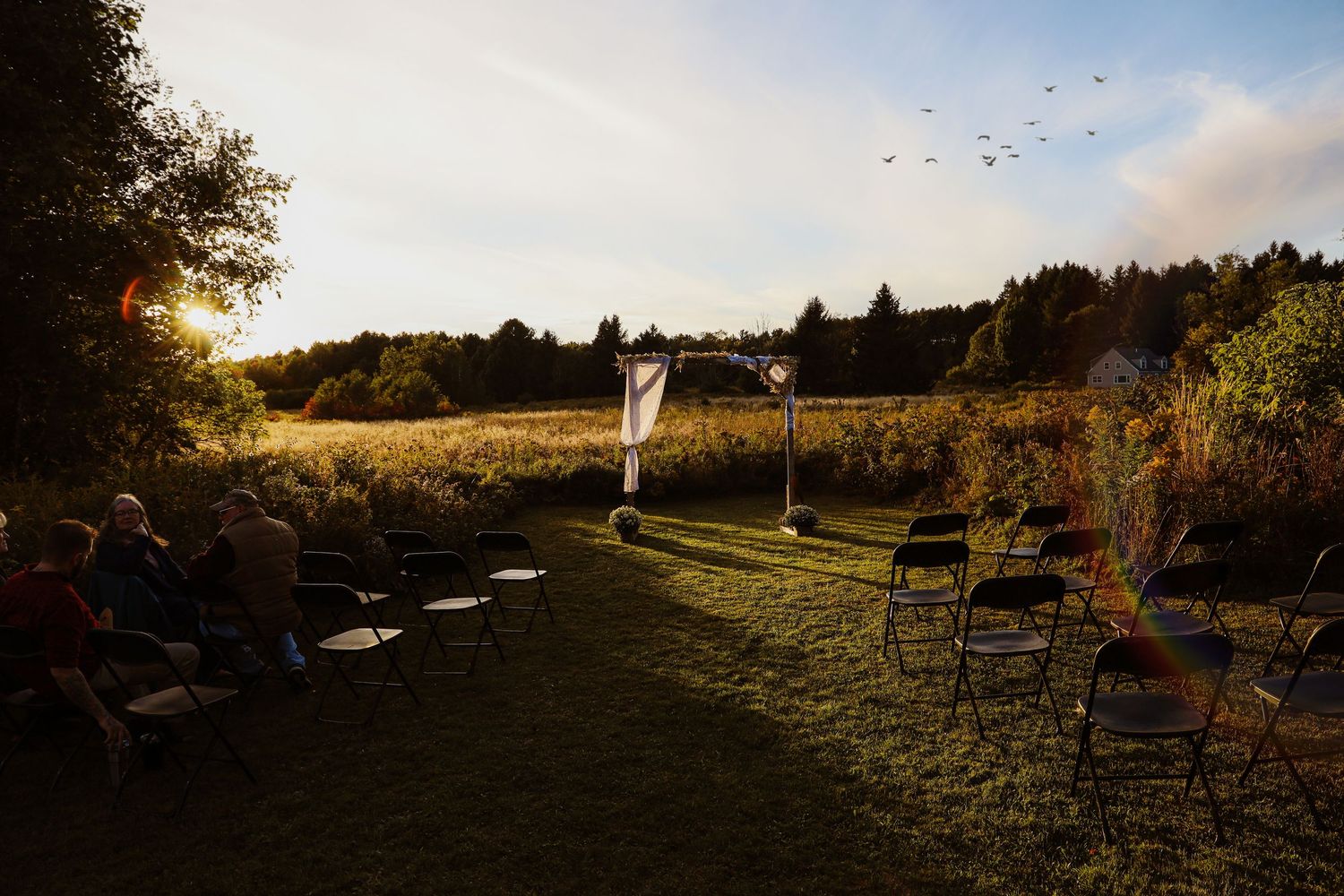 Outdoor wedding ceremony setup in a golden field at sunset with chairs and guests.
