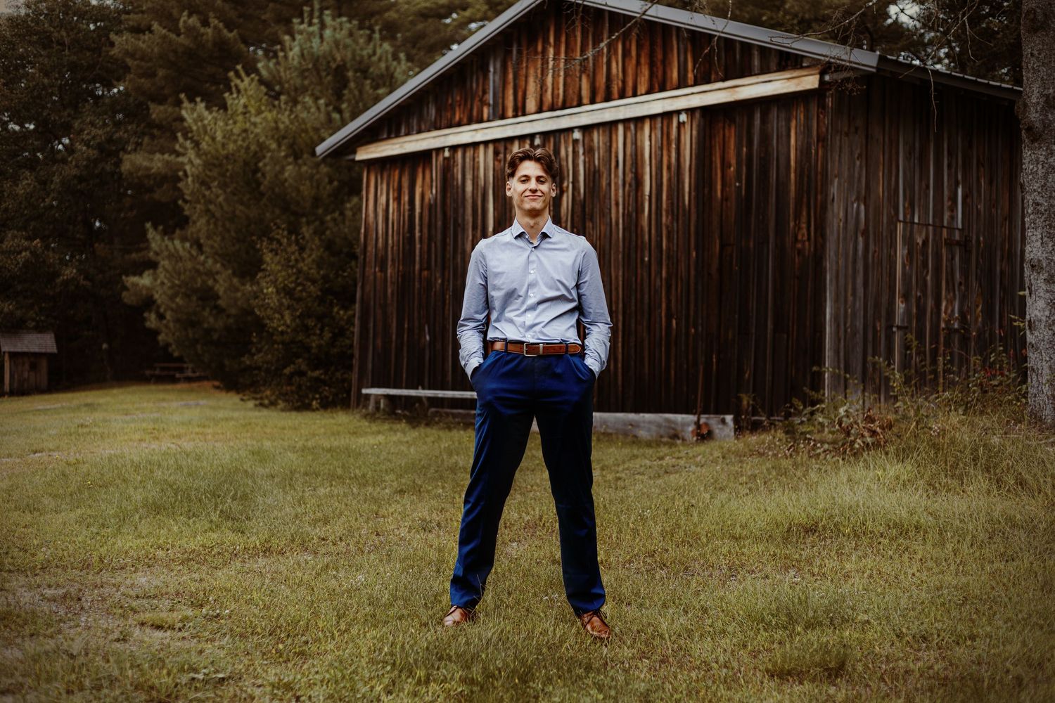 A person stands in business casual attire in front of a rustic wooden barn.