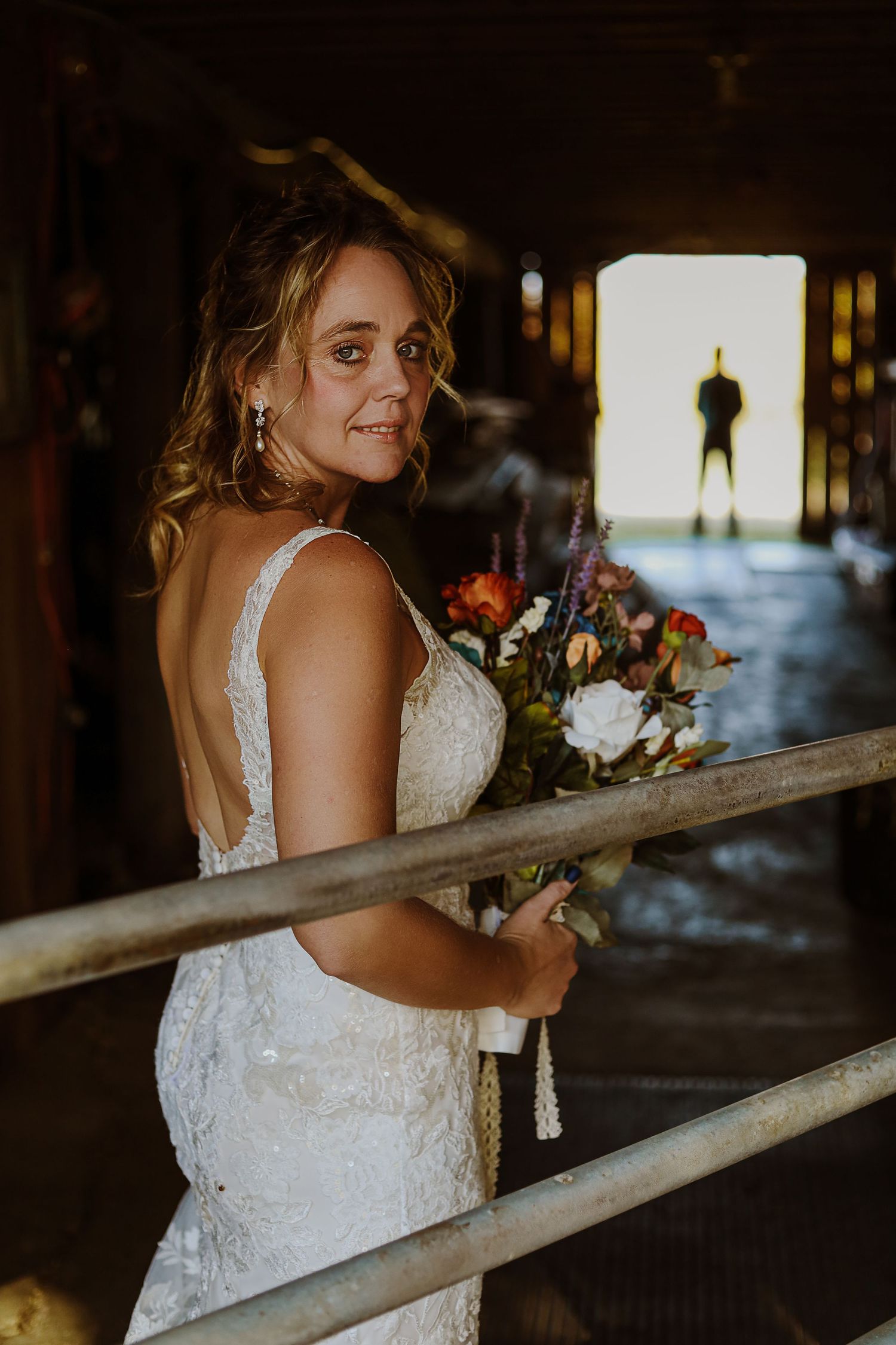 Bride holding autumn bouquet while standing near rustic railing with figure silhouetted in background.