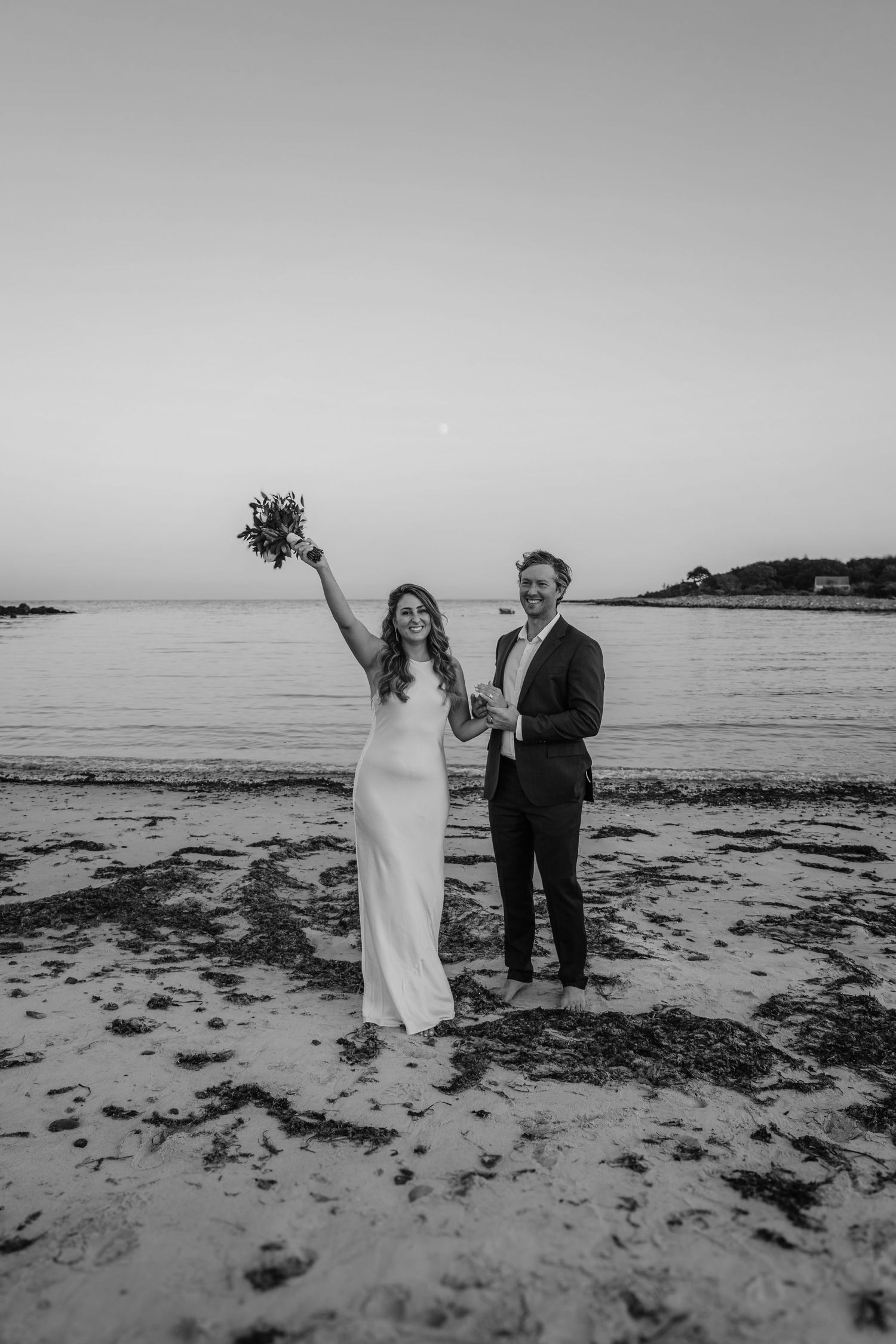 A couple celebrates on a beach at dusk holding a bouquet aloft in celebration.