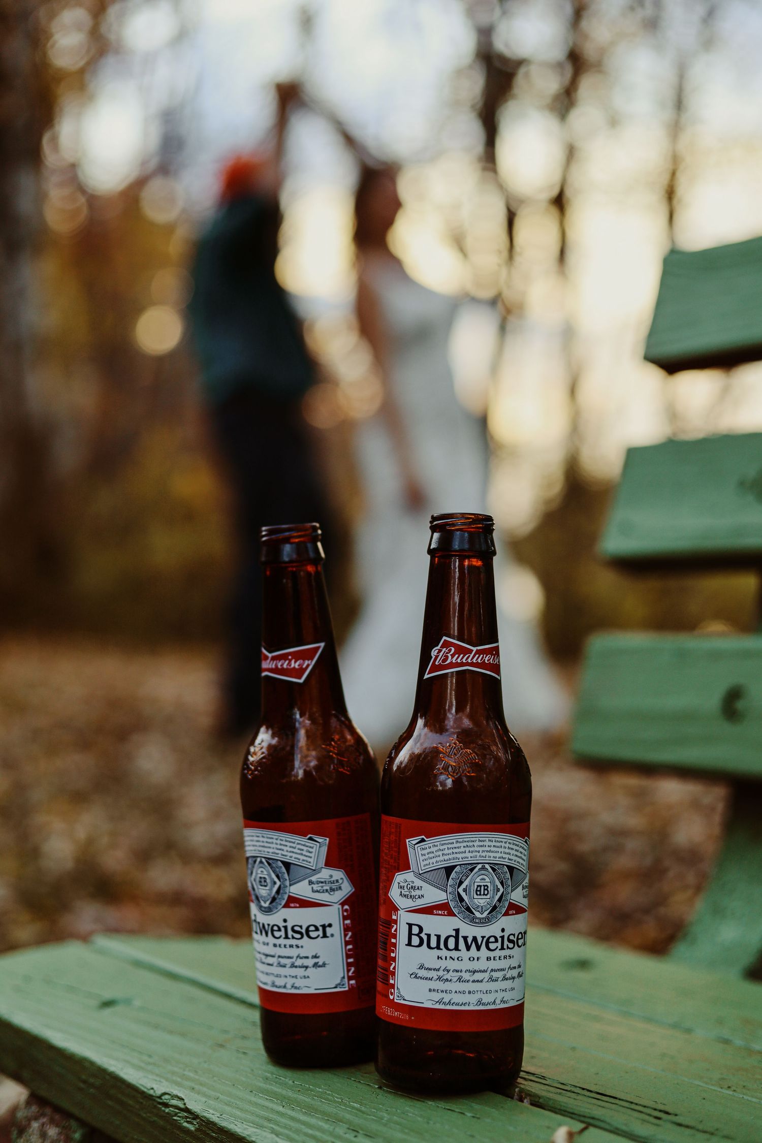 Two Budweiser beer bottles sitting on a green wooden surface with blurred outdoor background.