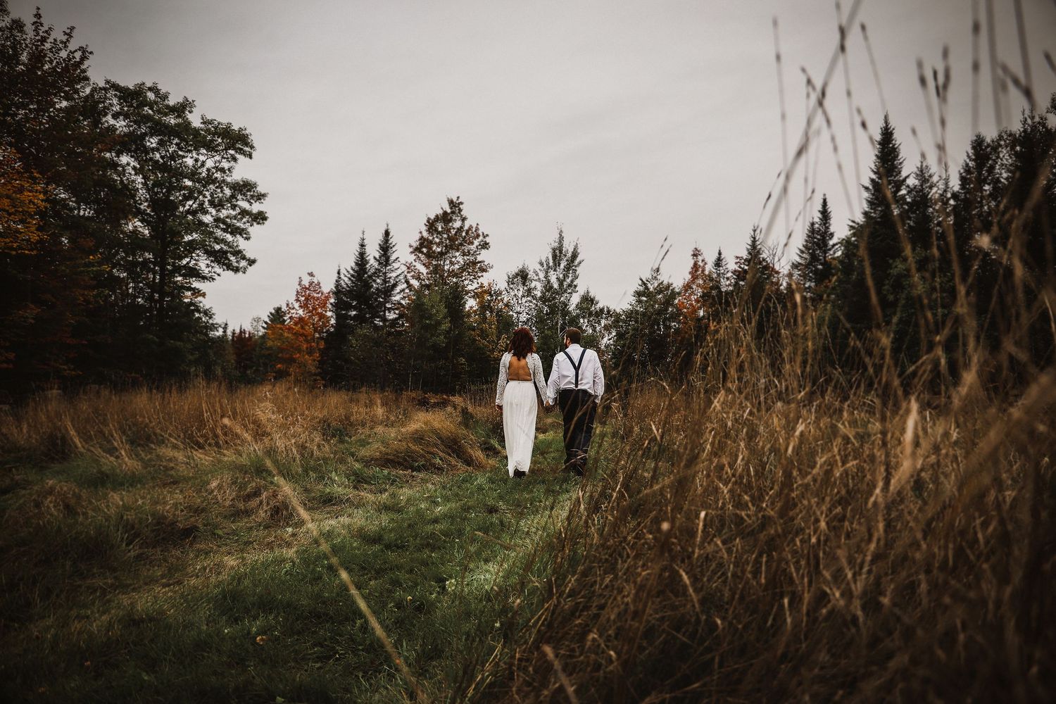 Couple walks through tall grass on forest path during autumn wedding.