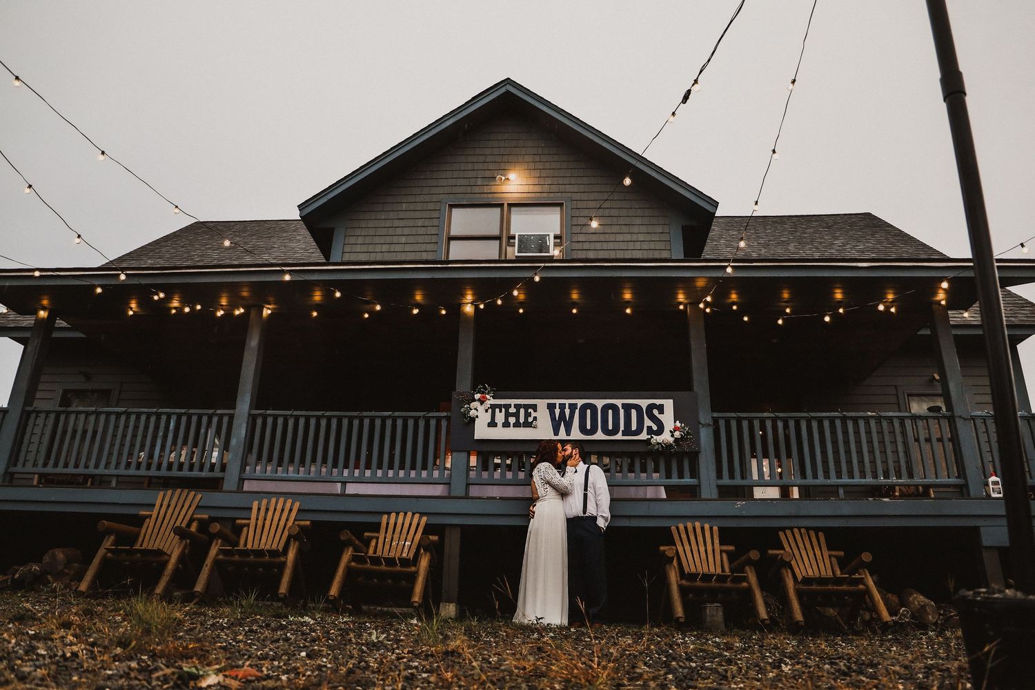 A rustic venue called 'The Woods' with string lights and wooden chairs on the front porch at dusk.