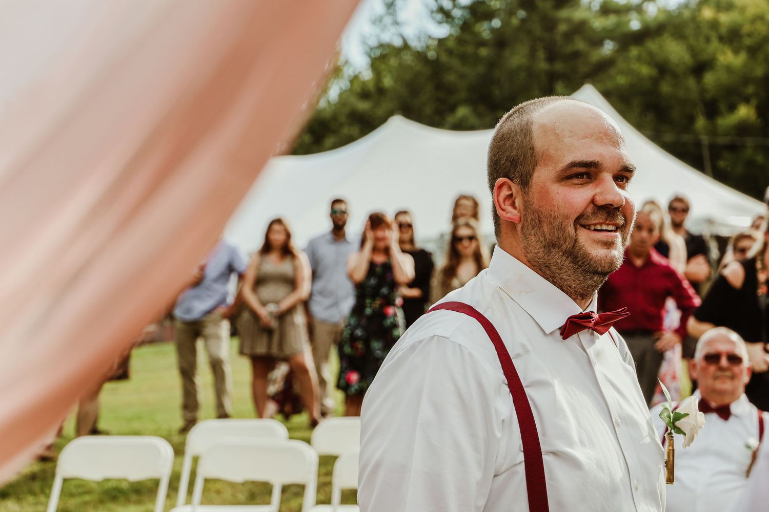 Wedding attendee in white shirt and suspenders smiling at outdoor ceremony.