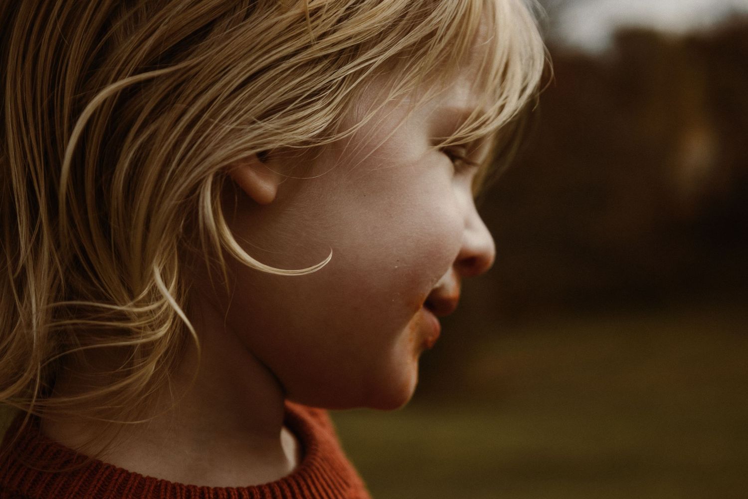 A young child in a red sweater looks to the side with a soft smile while windswept blonde hair frames their face.