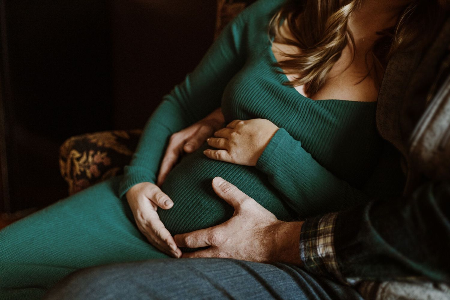 Intimate maternity portrait featuring hands cradling belly in deep emerald green sweater.