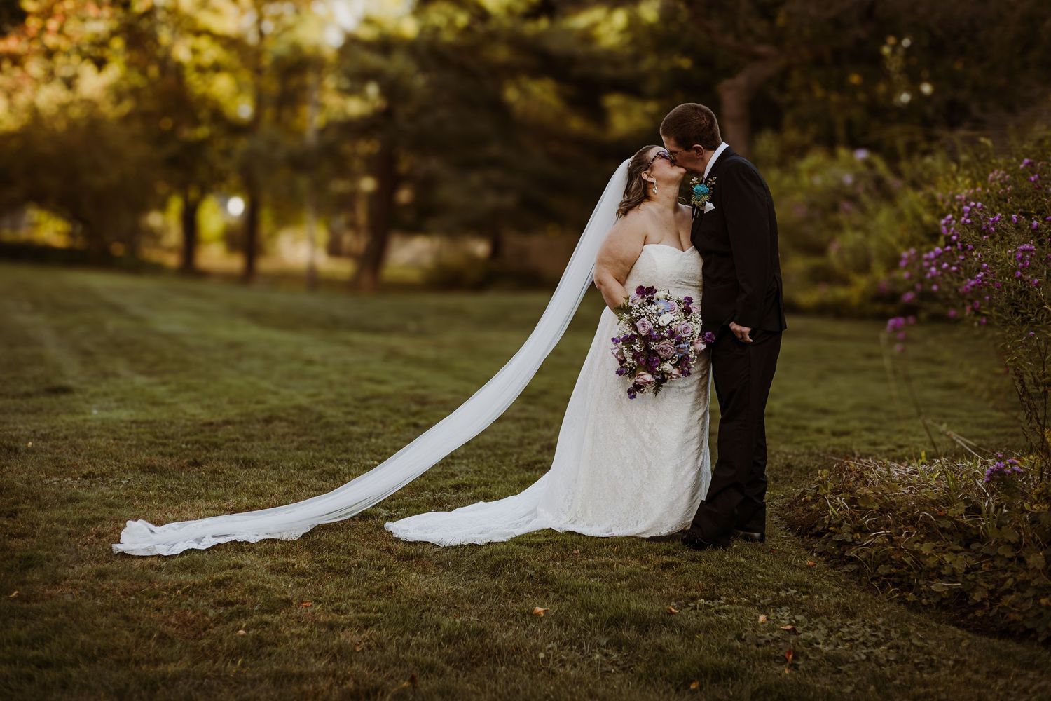 Newlyweds embrace on grassy hillside with dramatic long white veil trailing behind.