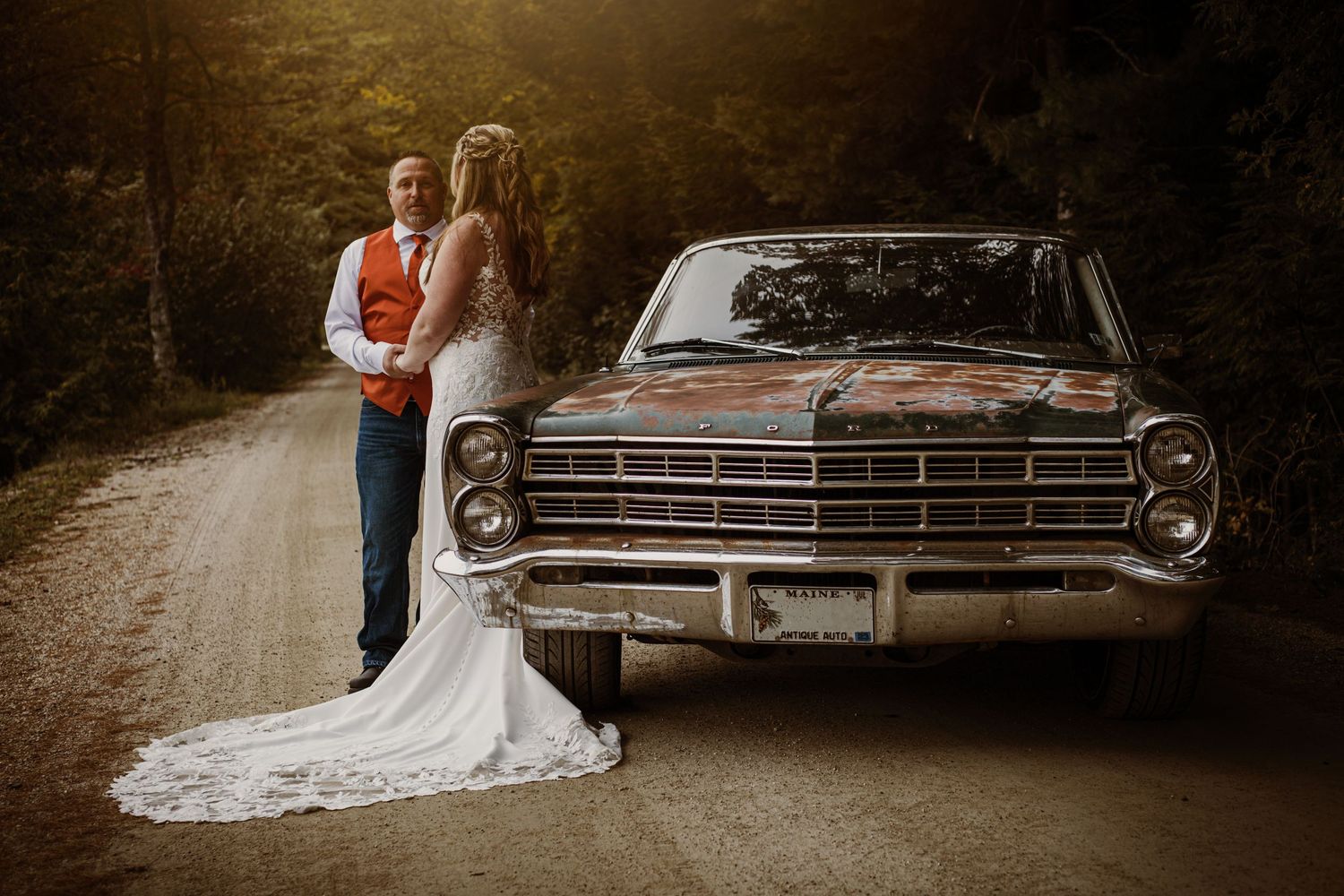 A vintage Ford Galaxie classic car serves as backdrop for a wedding portrait on a forest road.