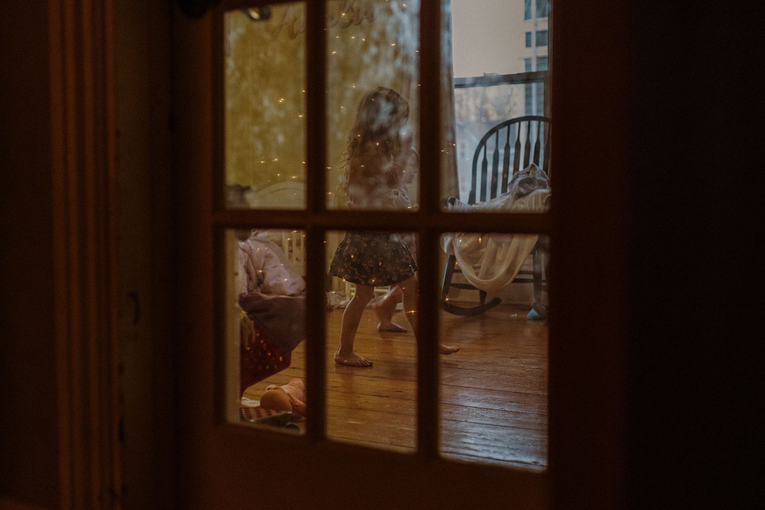 Looking through a paned window into a dimly lit dining room with wooden floors and chairs visible inside.