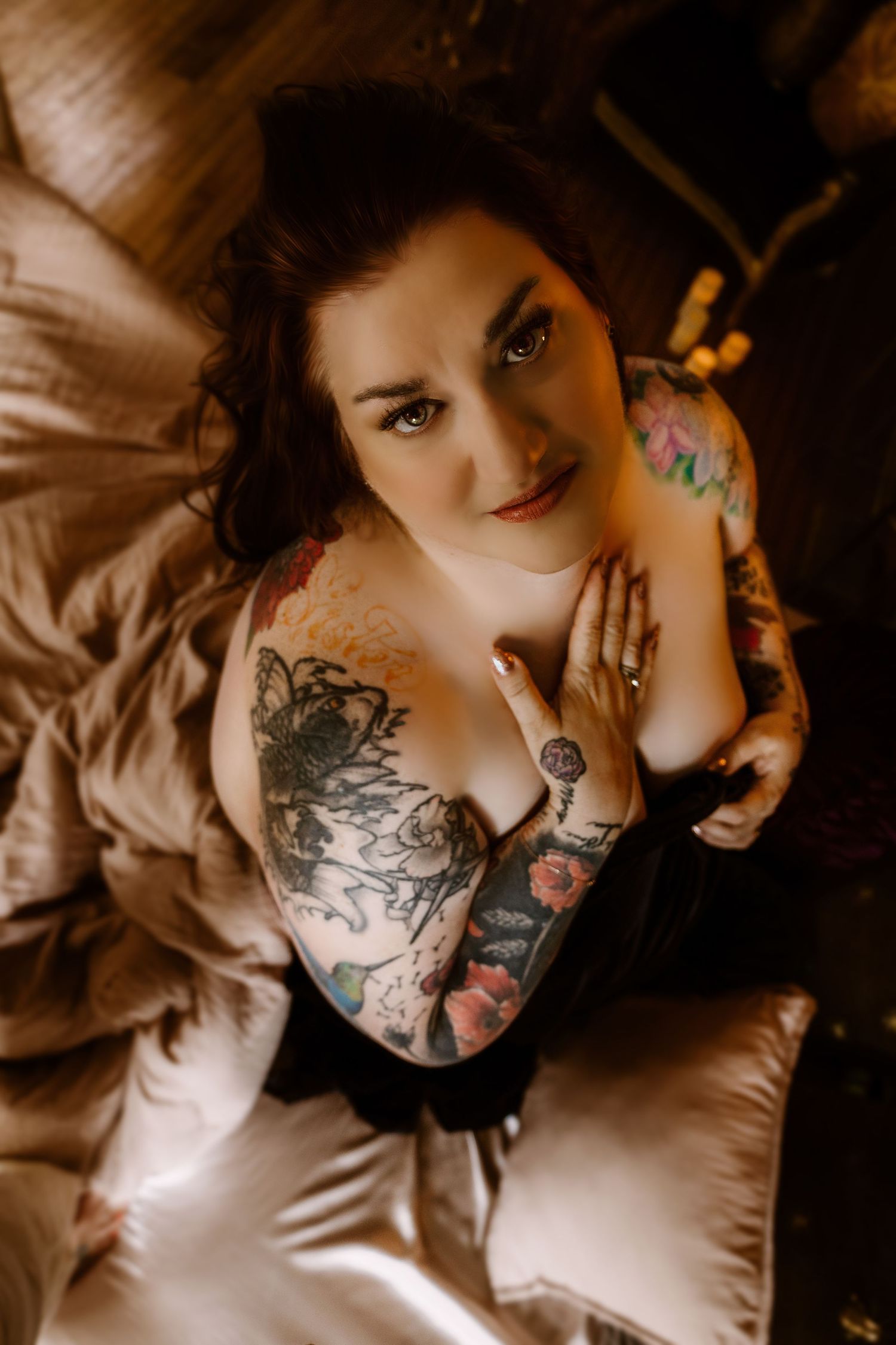 Overhead portrait shot with dramatic lighting showing intricate black tattoo artwork and dark lingerie on satin sheets.
