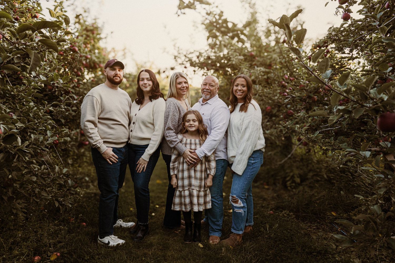 A group in casual attire poses together outdoors against a backdrop of green foliage at sunset.