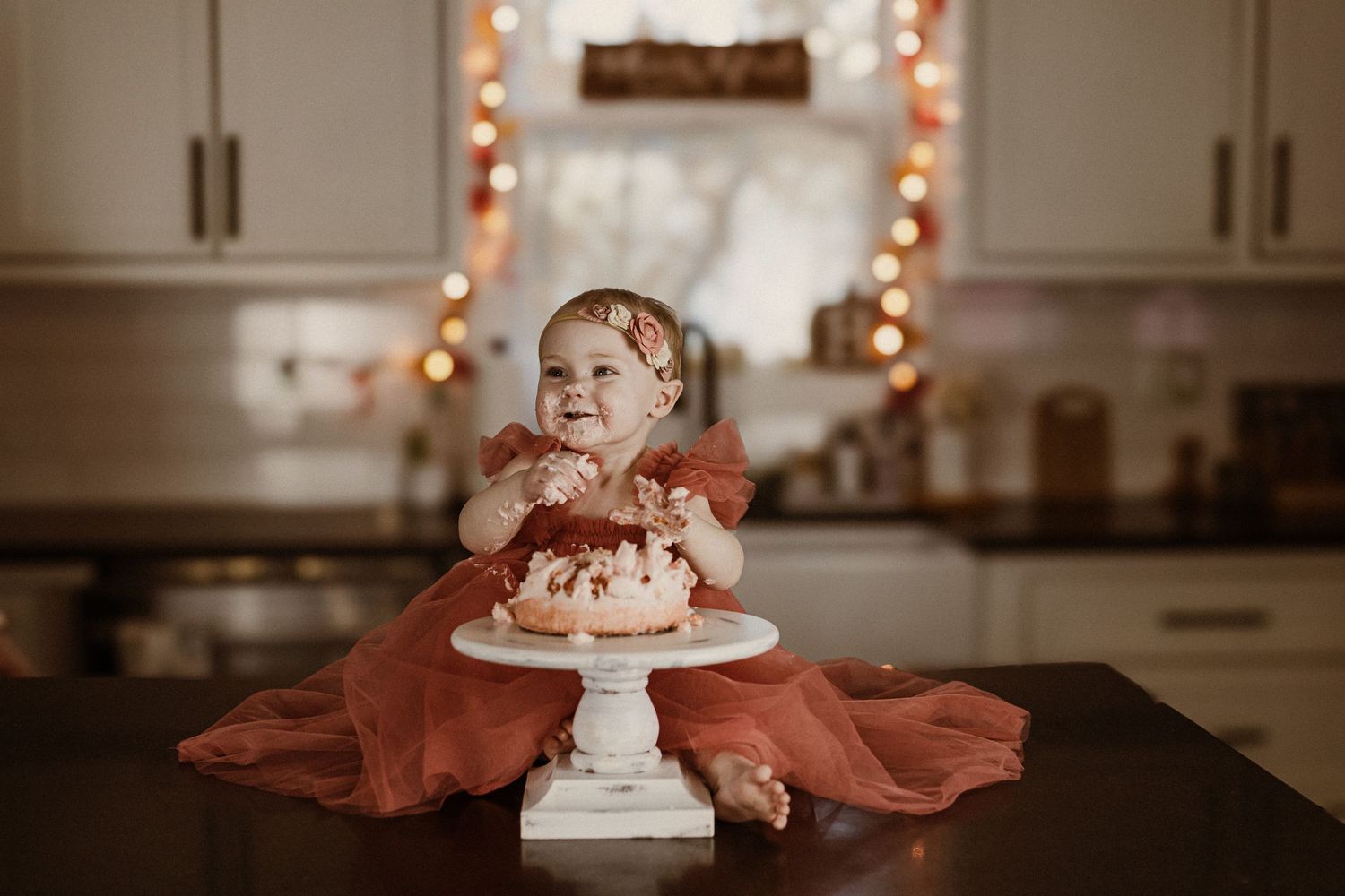 A small child in red dress sits at table with birthday cake and twinkle lights.
