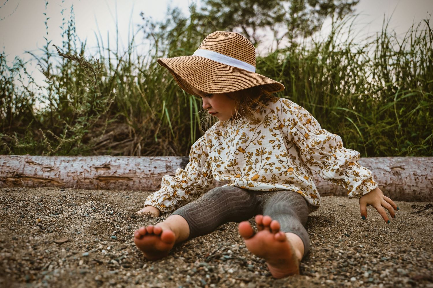 A child in a floral dress and sun hat sits on rocky ground with bare feet extended in warm sunlight.