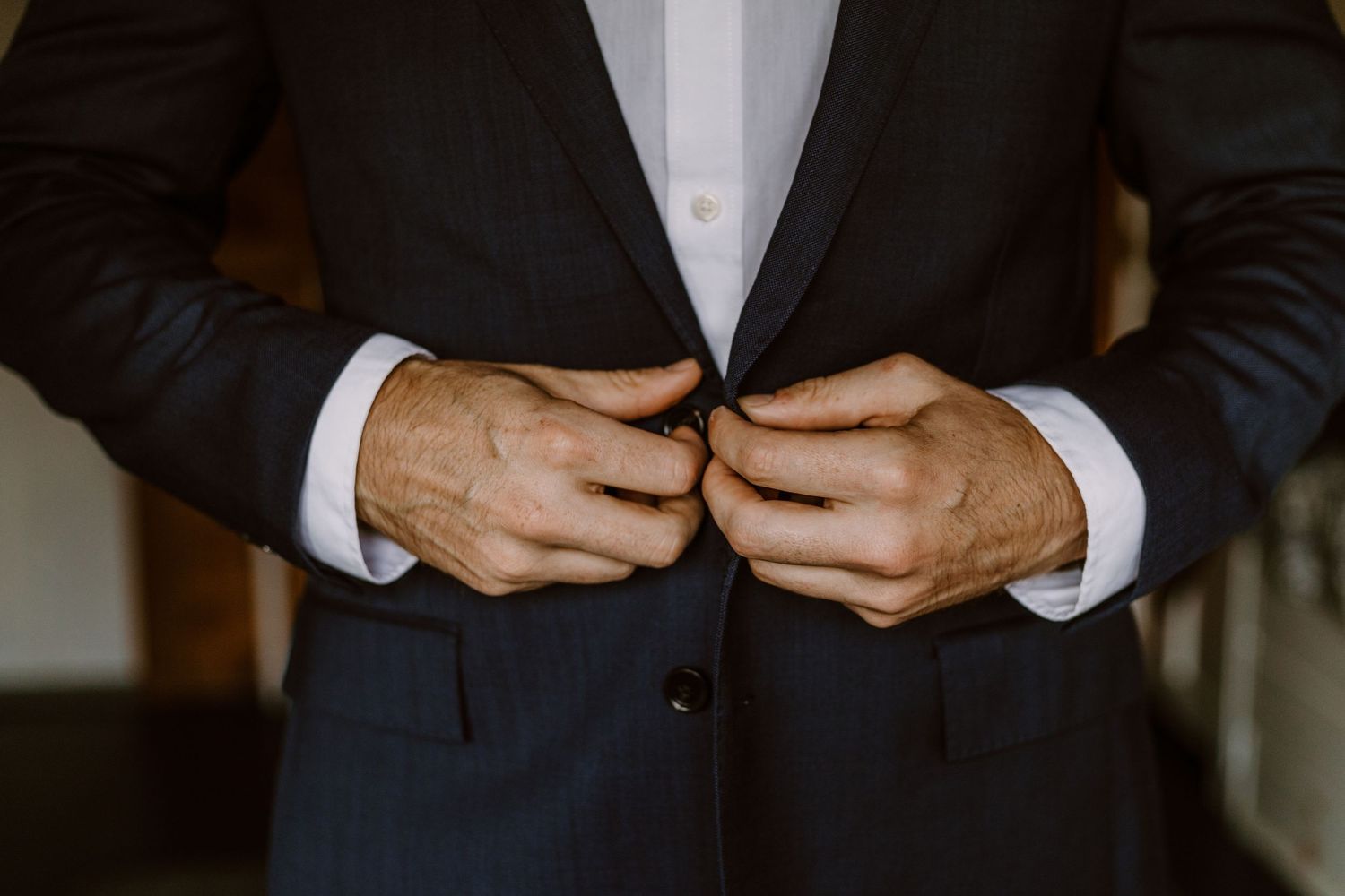 Close-up of hands buttoning a classic black suit jacket with white dress shirt cuffs visible.
