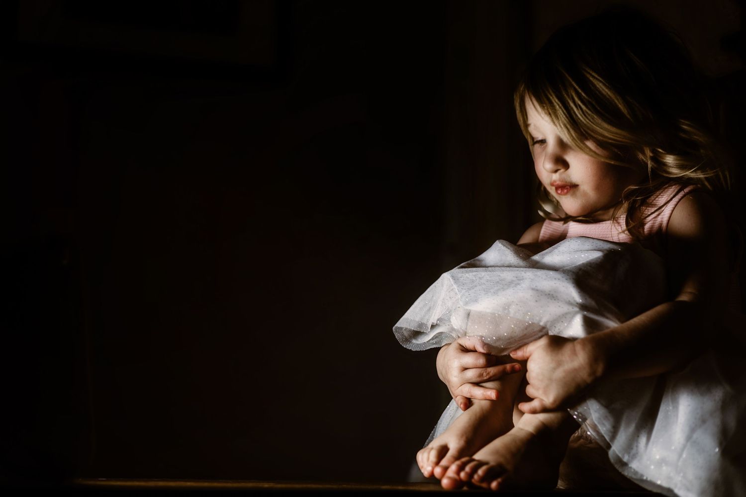 Dramatic moody low-key portrait of small child in white dress sitting in shadow with light falling across dark scene.