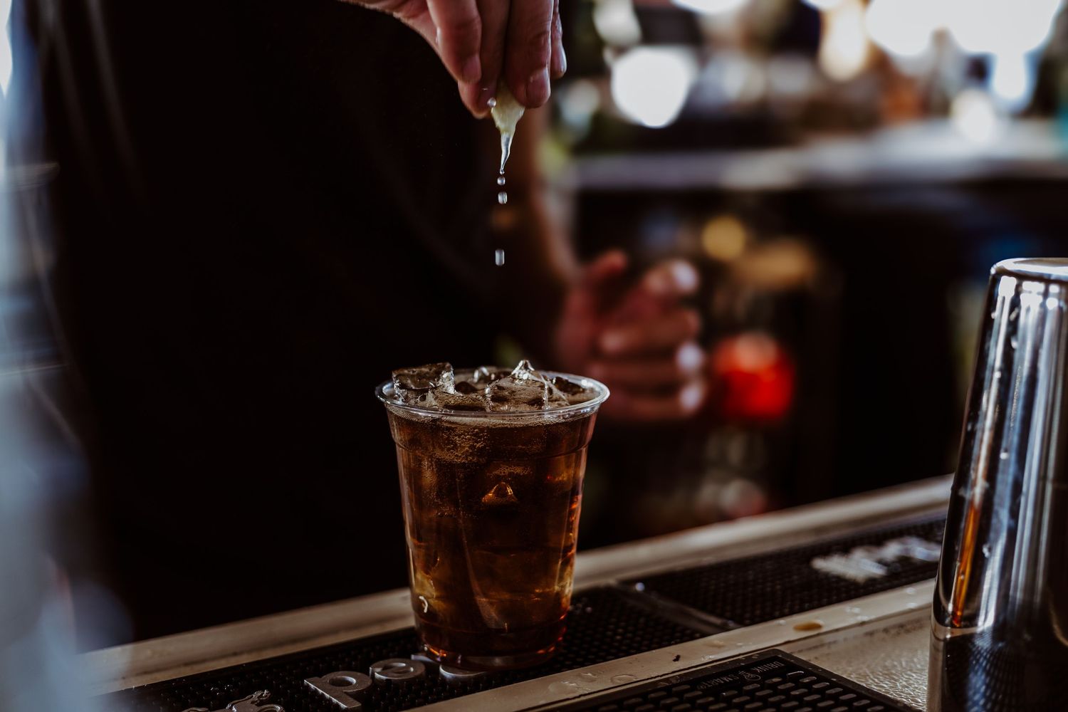 Bartender pouring a dark beverage into a plastic cup with ice at a dimly lit bar counter.