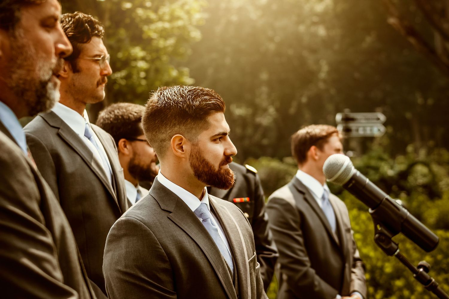 Groomsmen in dark suits stand together during outdoor wedding ceremony.