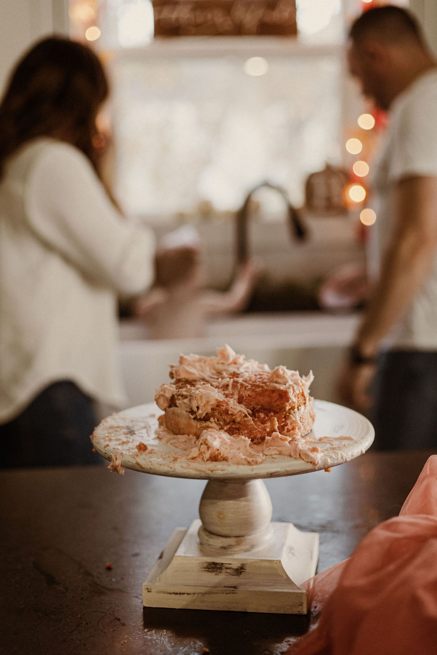 A rustic cake stand displays a textured dessert while people gather in a softly lit background.