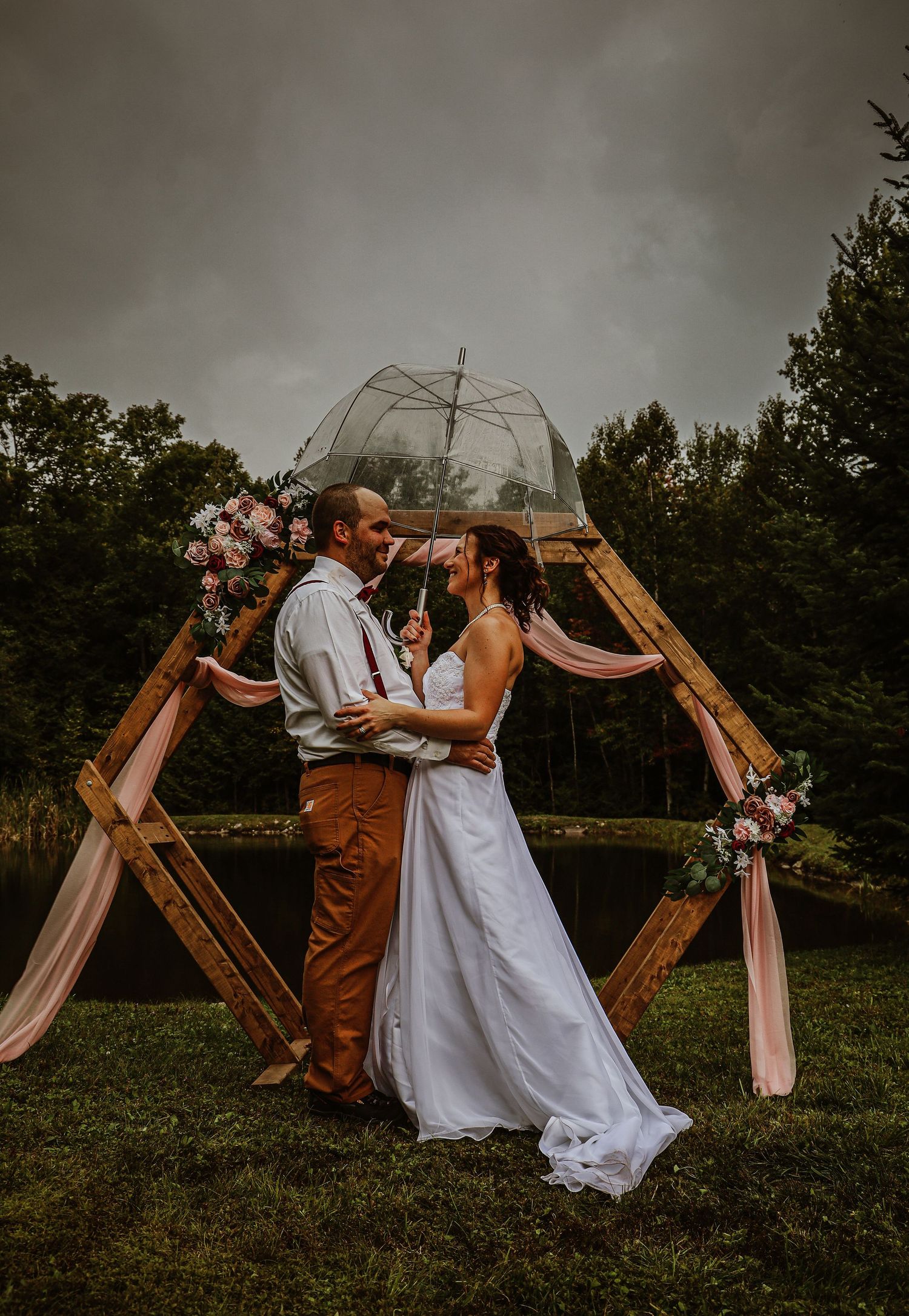 Couple embraces under wooden arch with floral decorations at moody outdoor wedding.