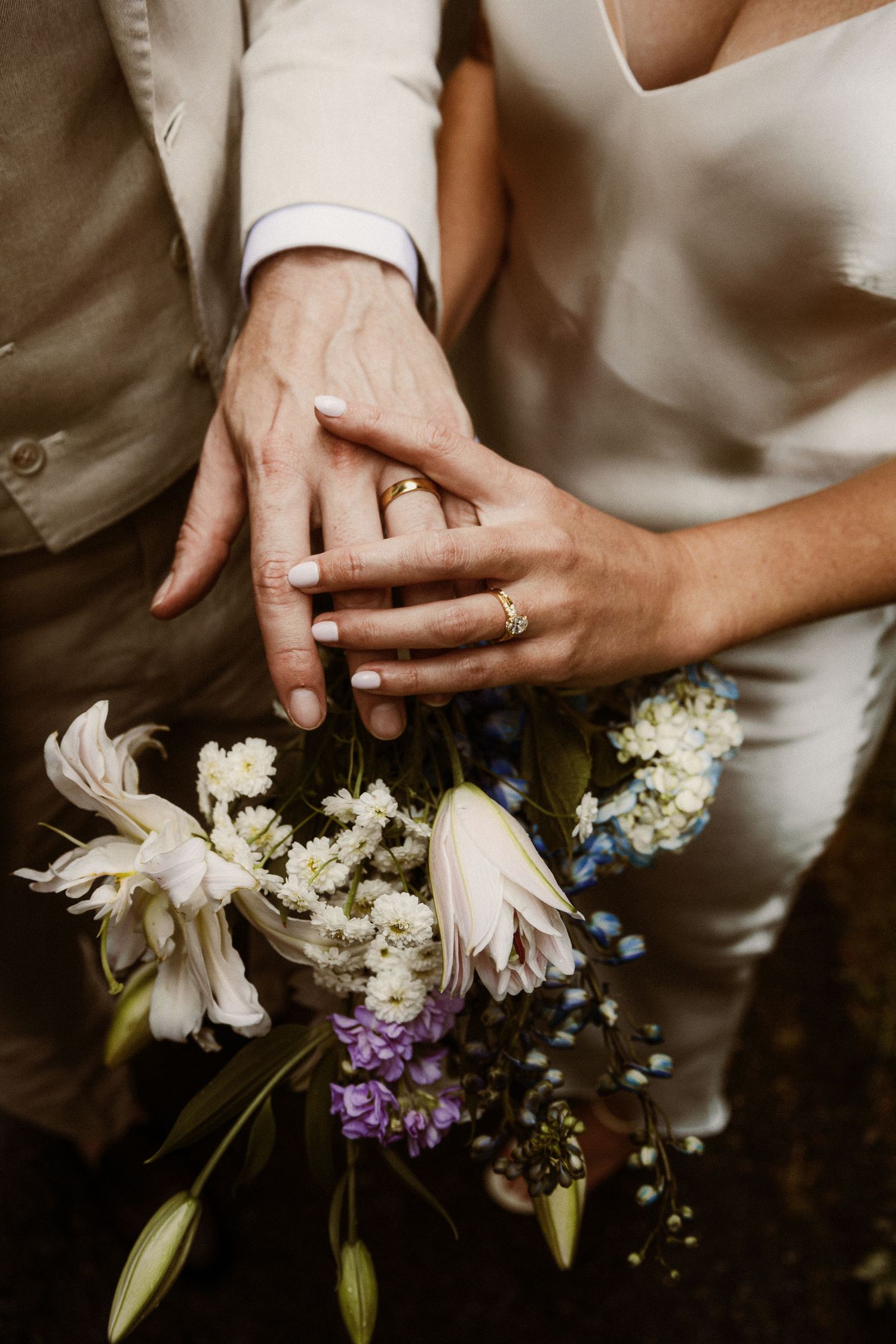 Close-up of wedding rings and hands with delicate white and purple flowers.