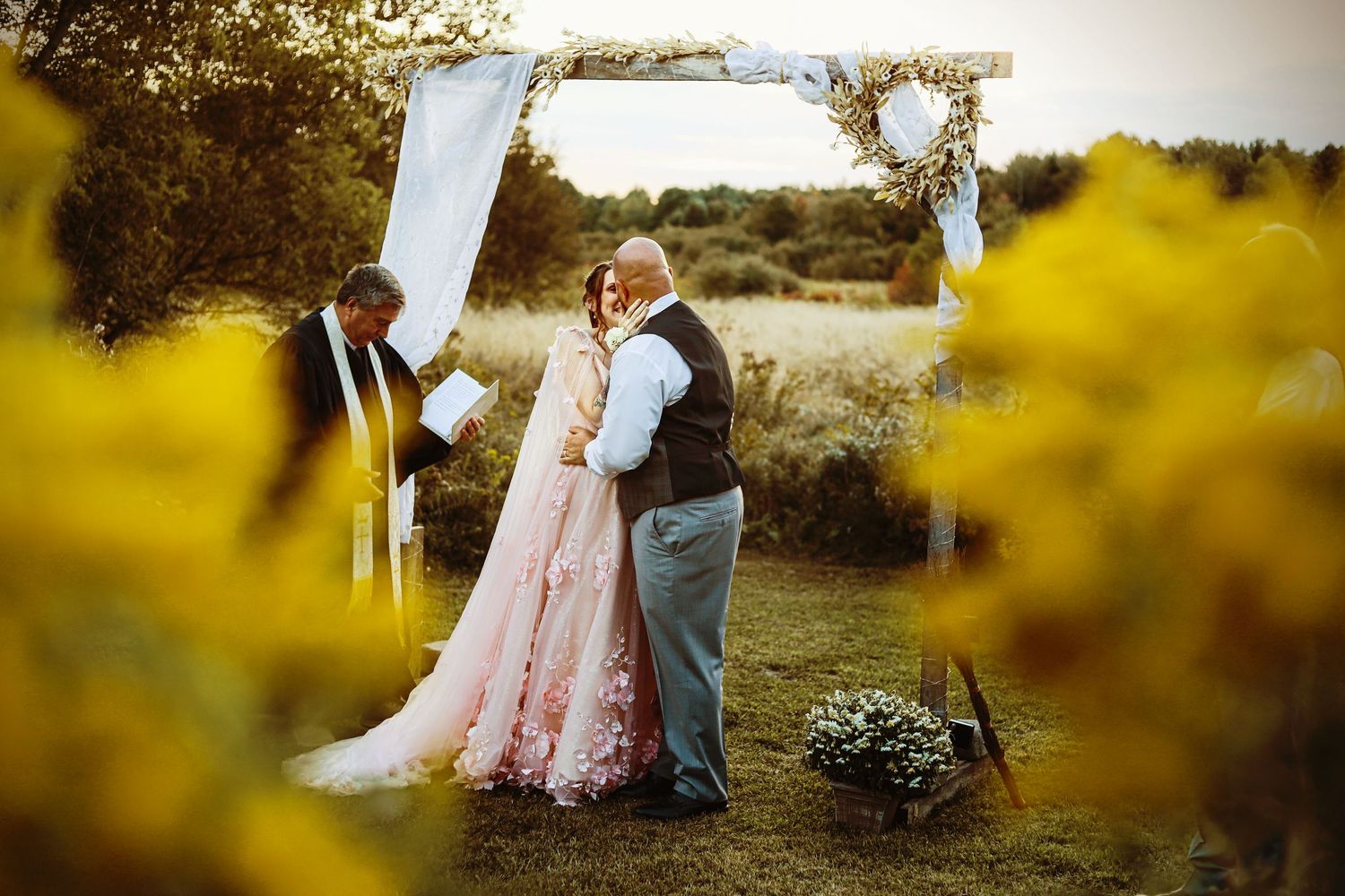 Wedding couple embraces under a floral arch with yellow flowers in foreground.