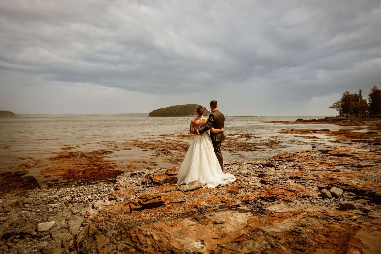 Newlyweds embrace on rocky coastal shore under dramatic cloudy sky.