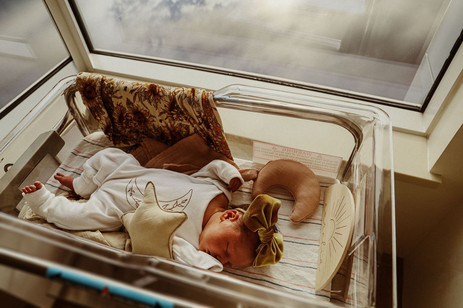 Overhead view of infant resting in hospital bassinet beneath window.