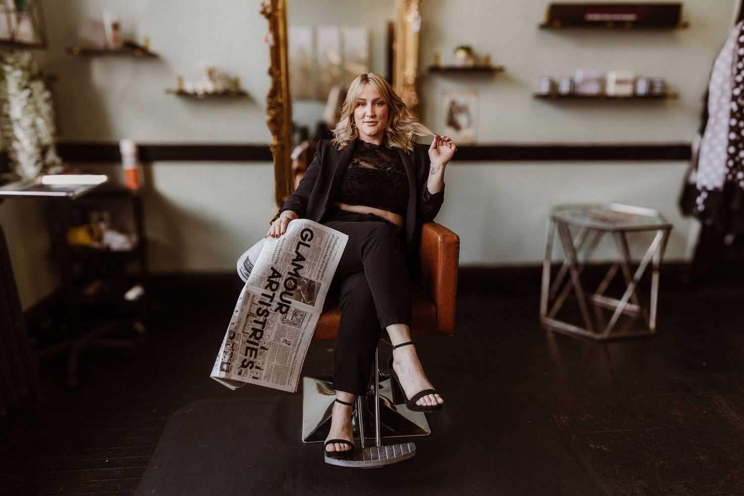 A person in black attire sits casually in a chair holding a newspaper in a rustic room.