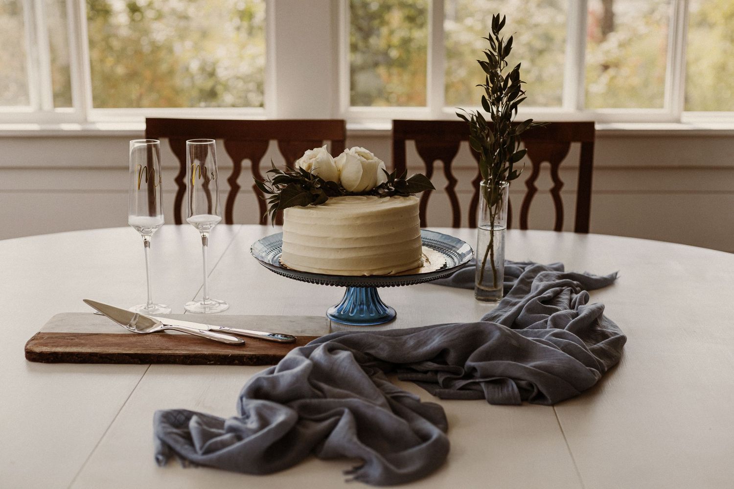 White cake on blue pedestal stand with decorative foliage and wine glasses on rustic table setting.