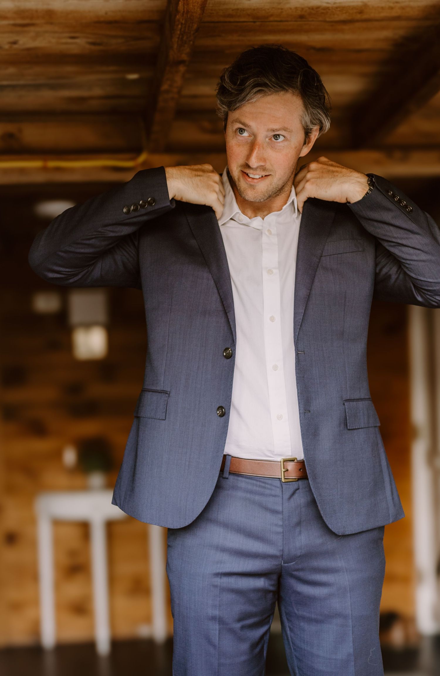 A suited figure adjusts their collar while standing in a rustic wood interior.