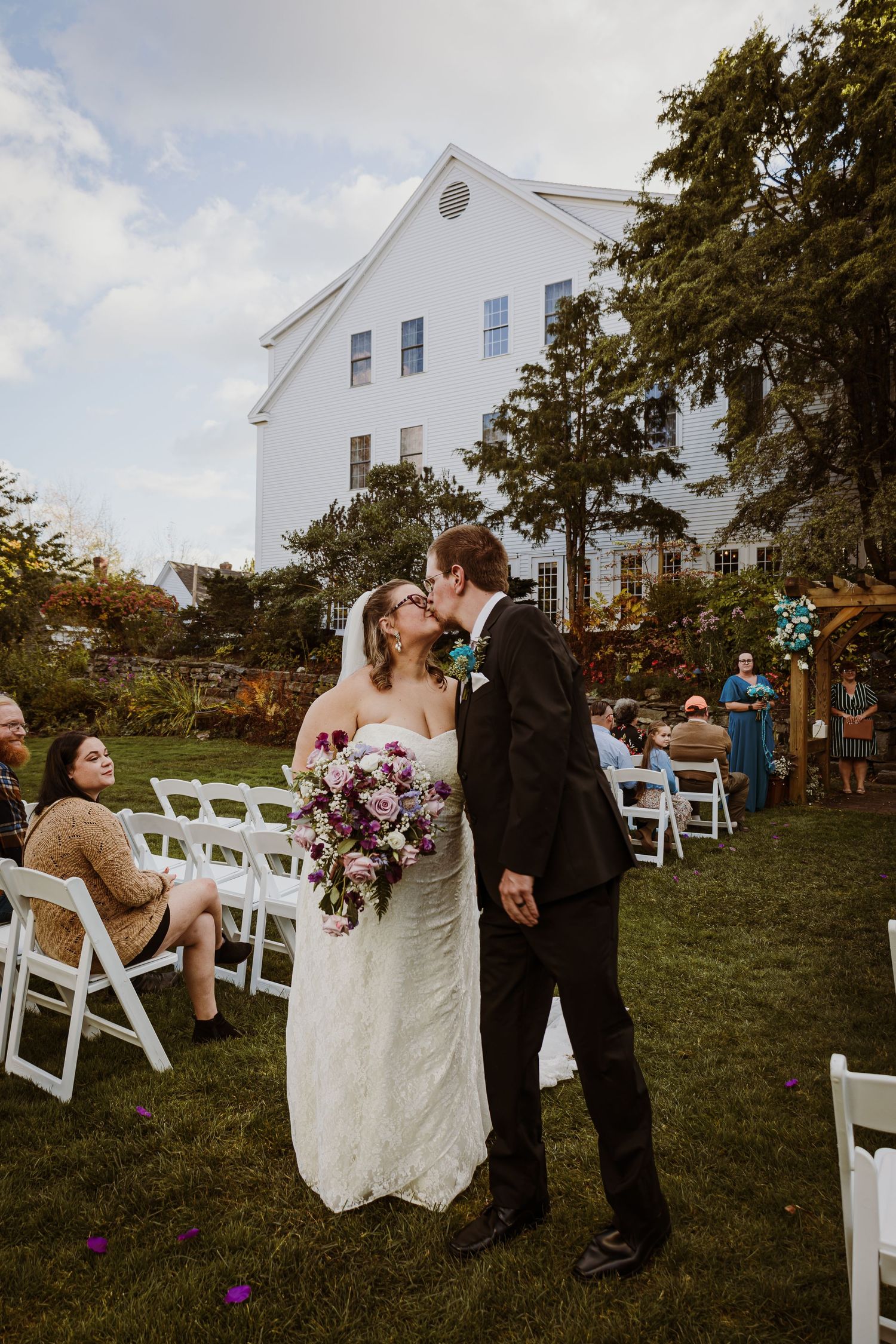 Wedding couple sharing a kiss outside a white colonial-style building with guests seated on lawn chairs.