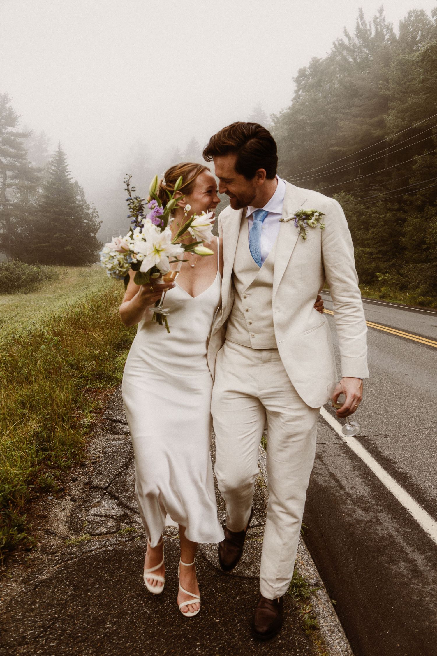 A romantic wedding photo on a foggy mountain road with the couple wearing matching cream-colored attire and holding flowers.