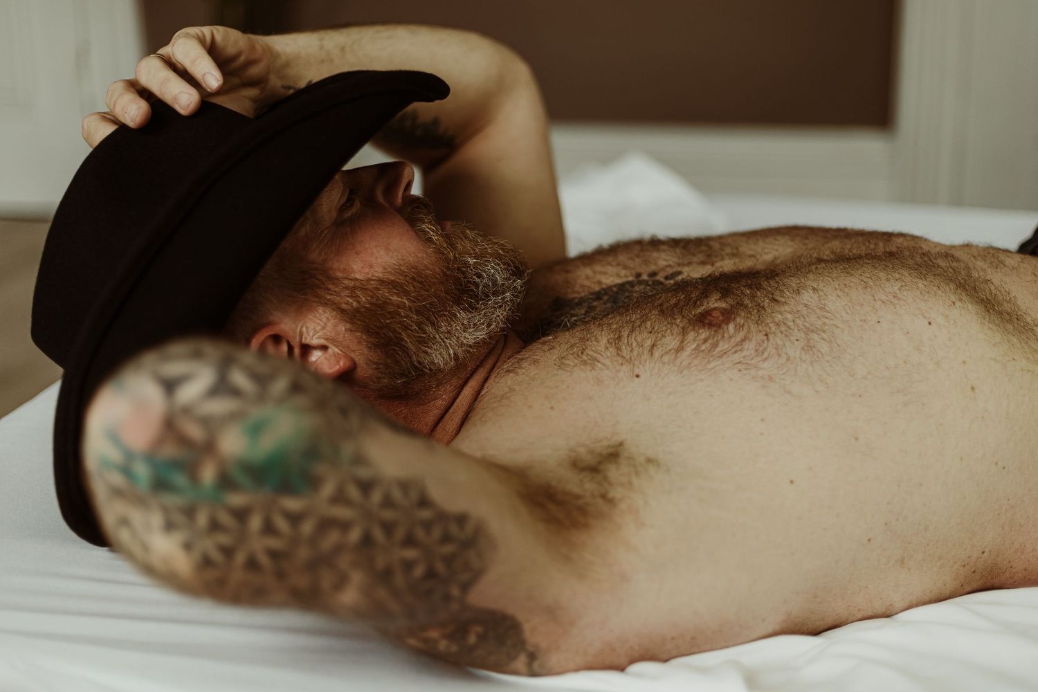 Shirtless person with geometric tattoos and a black hat lies on white bedding in moody lighting.
